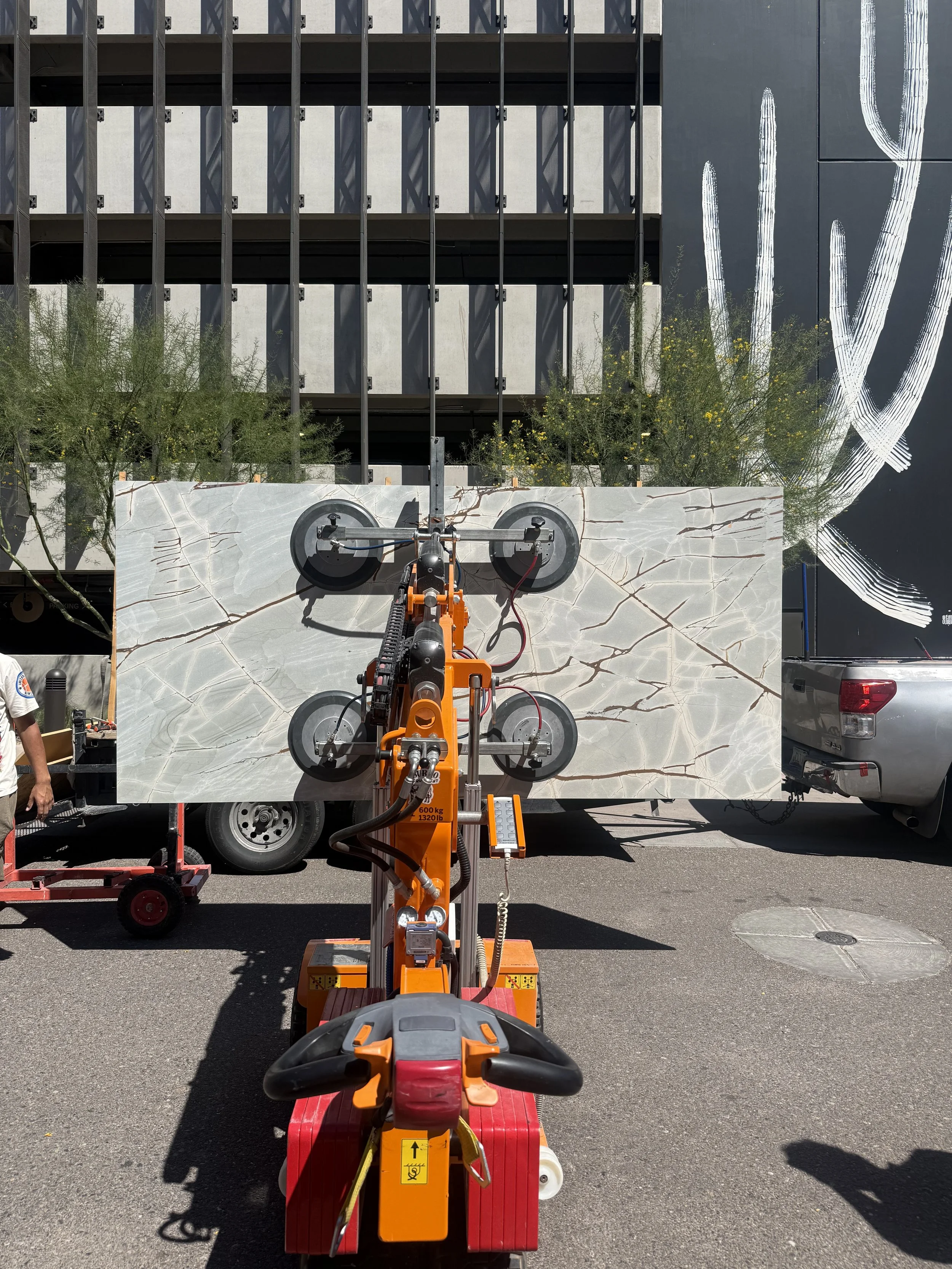 Construction equipment lifting a large marble slab outside a building, with trees and a black wall with white graffiti in the background.