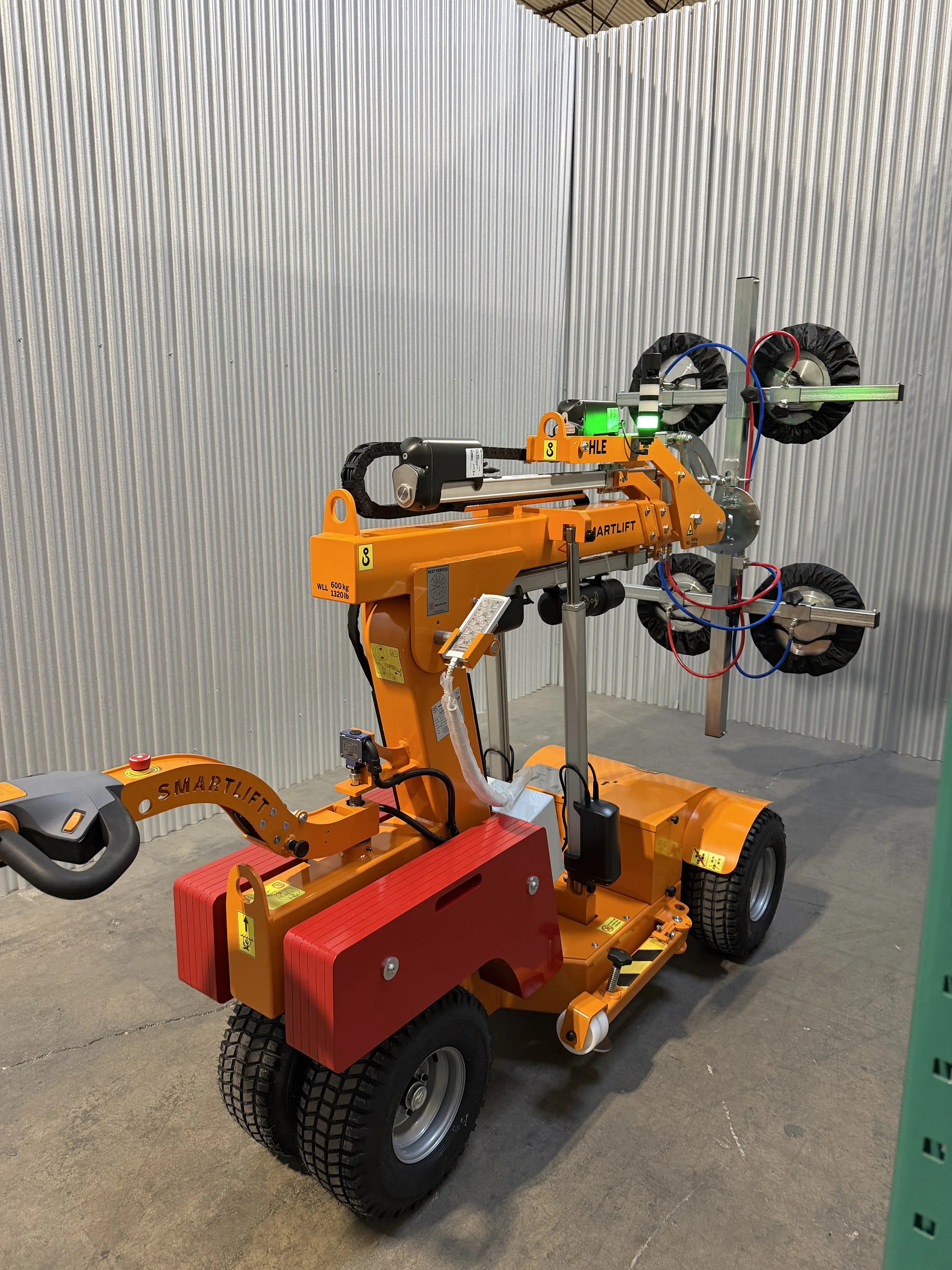 Small autonomous robot with orange frame and black wheels, equipped with mechanical arms, sensors, wiring, and a control panel in a warehouse with corrugated metal walls.