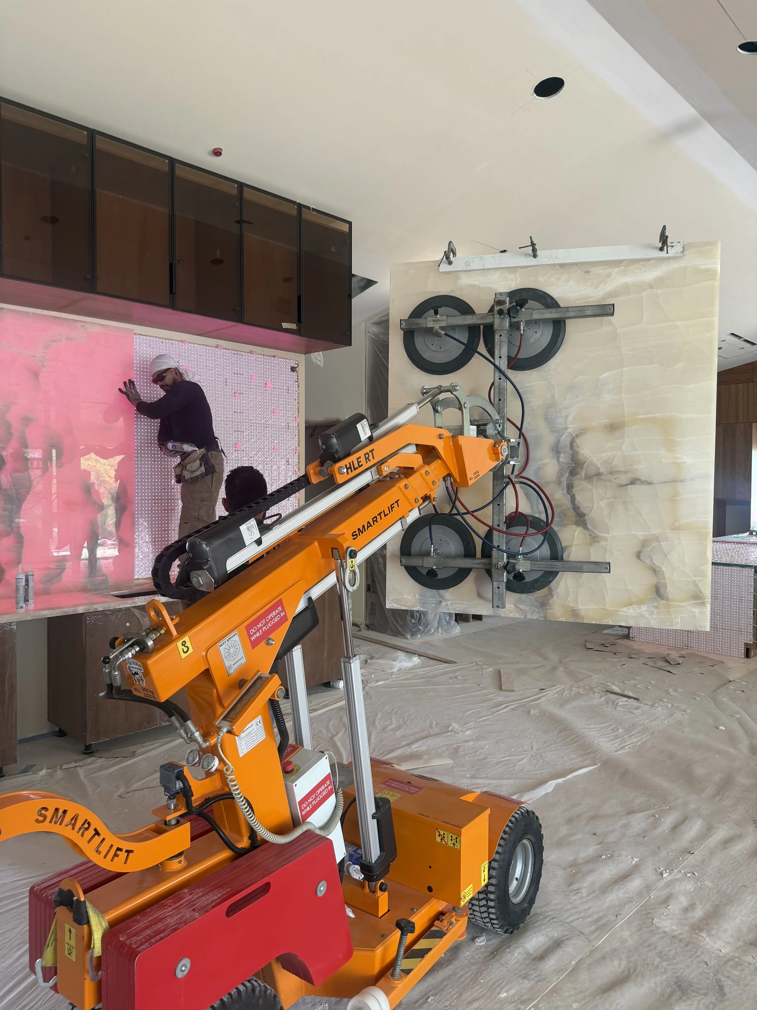 Construction worker installing or working on a feature wall with pink sheets, while a large marble slab is being moved or positioned with a specialized lift. The room is under renovation with protective covering on the floor.