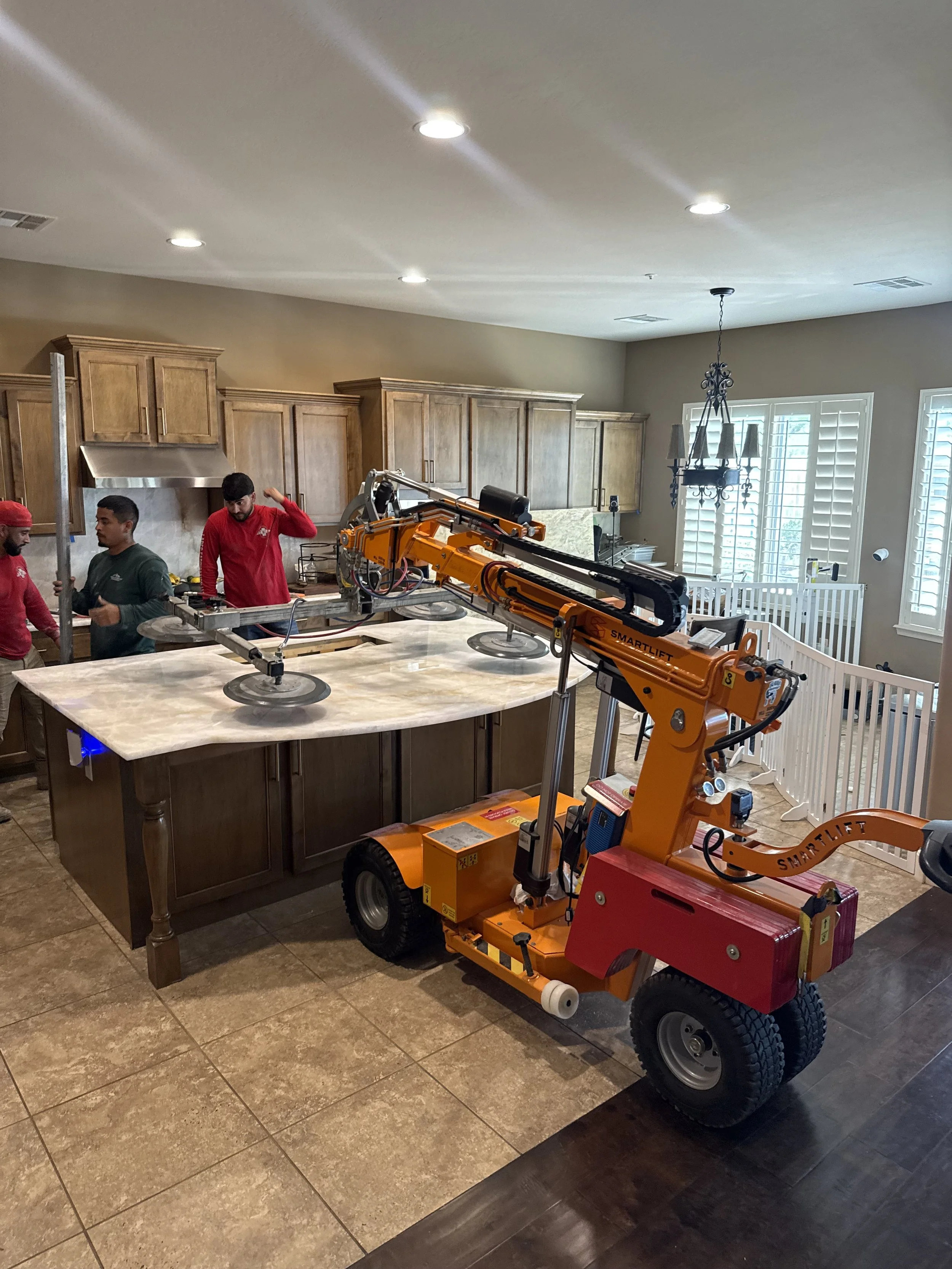 A robotic lift in a kitchen with wooden cabinets, marble countertops, and a tiled floor. Three workers are present, one wearing a red shirt, another in gray, and the third in red, all near the robotic lift which is positioned on the floor.