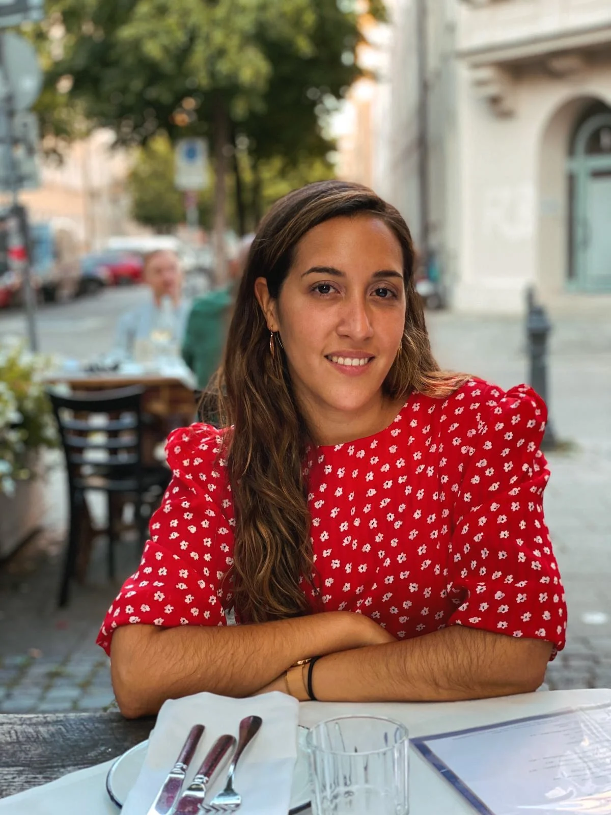 A young woman with long brown hair and hoop earrings, wearing a red dress with white floral pattern, smiling and sitting at an outdoor restaurant table with cutlery and a glass, in an urban setting with blurred background of pedestrians and buildings.