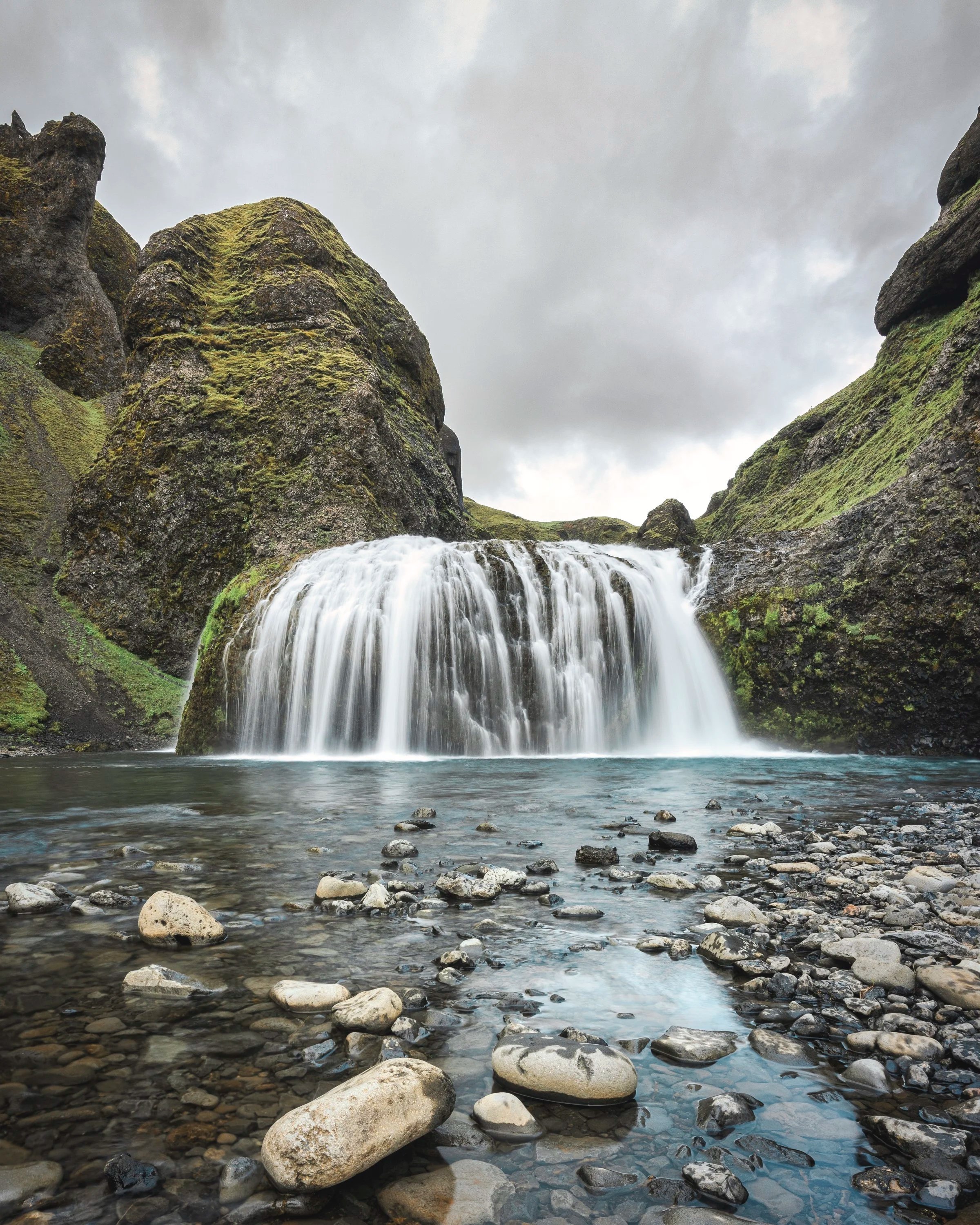 Ein Wasserfall, der in einem Tal mit grünen, moosbedeckten Felsen und einem Fluss mit glatten Steinen fließt, unter einem bewölkten Himmel.