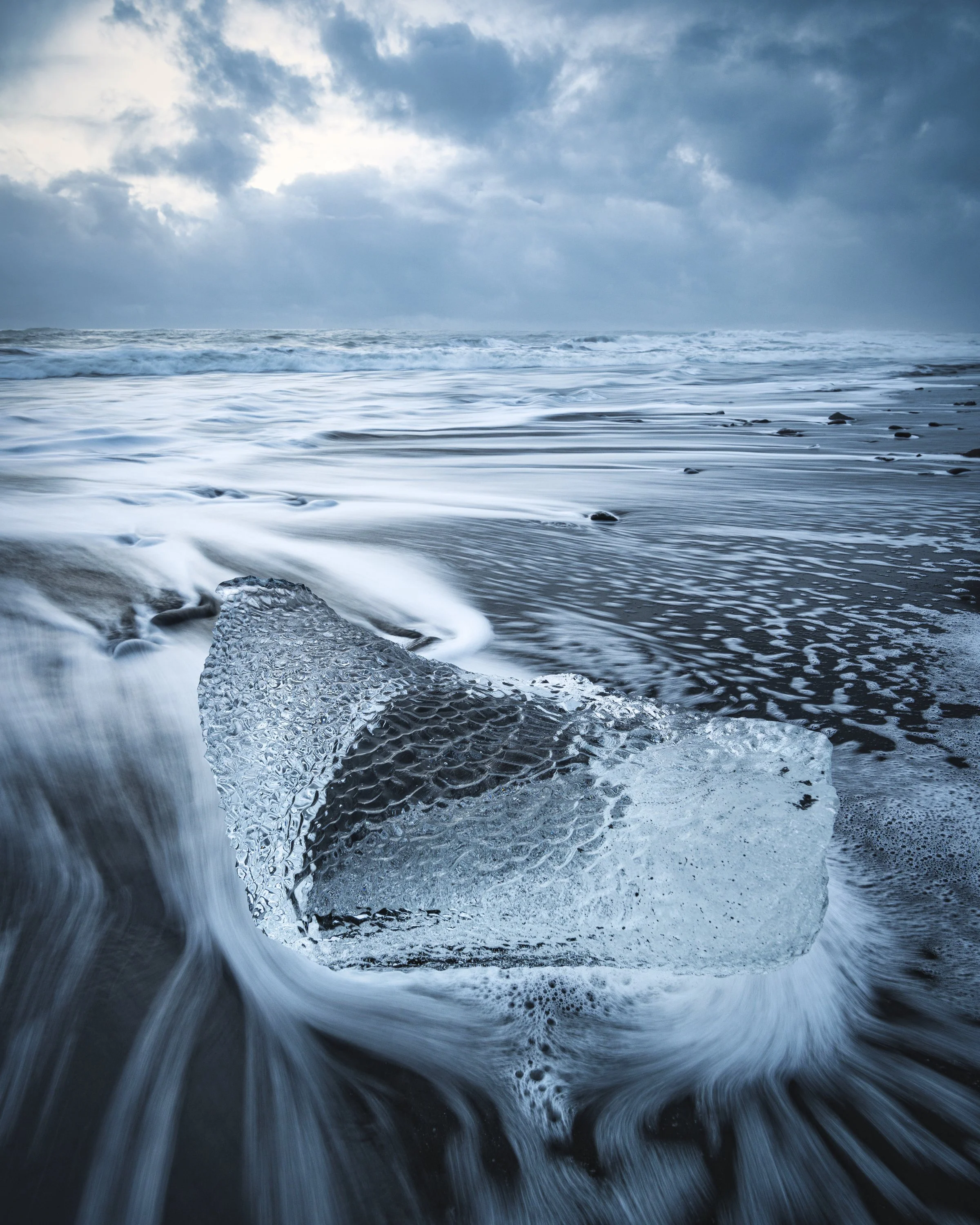 Eisstück am Strand von Island, das vom Wasser umspült wird, mit Ocean und bewölktem Himmel im Hintergrund.