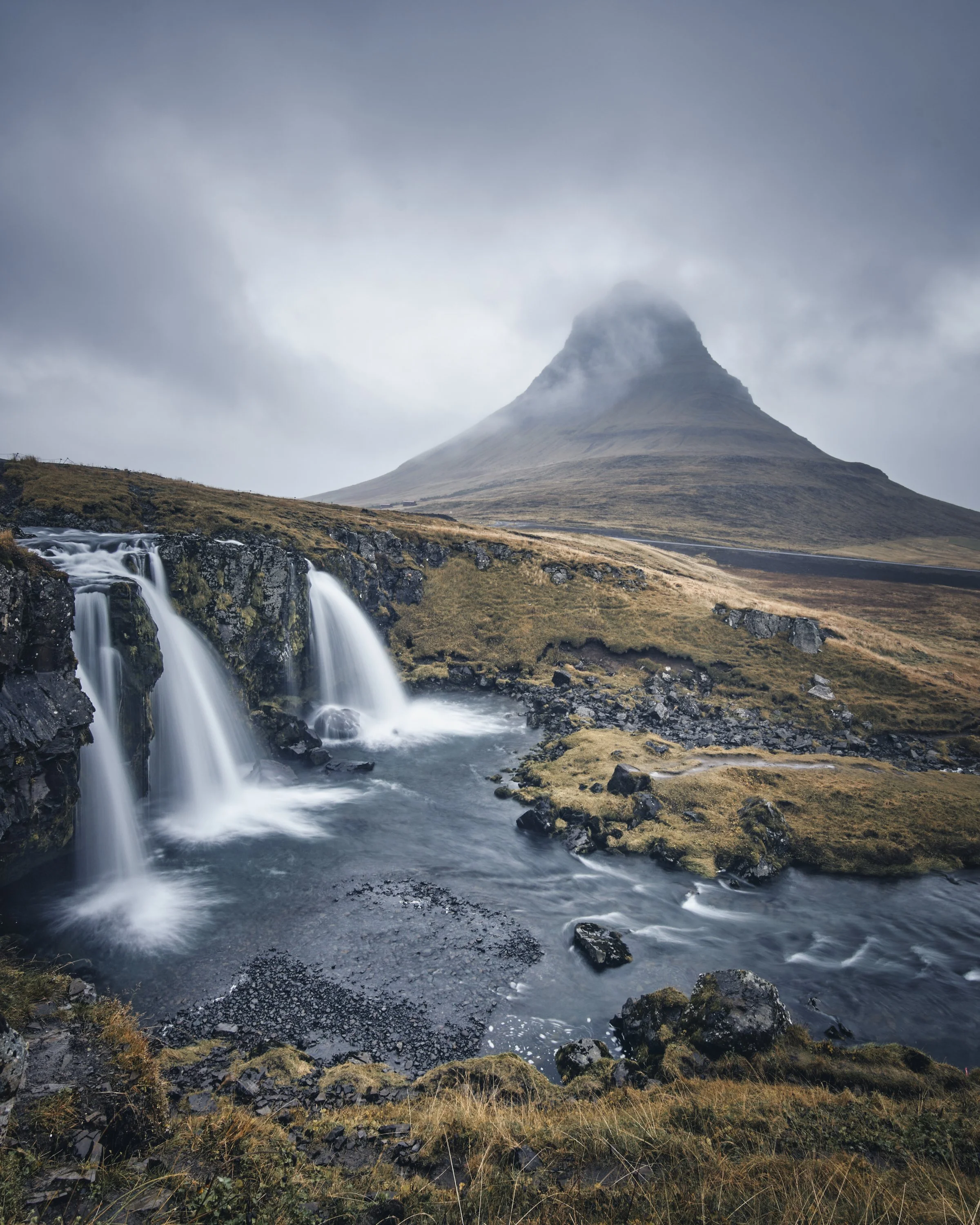 Ein nebliger Berg mit einem Wasserfall und einem Fluss im Vordergrund, umgeben von Gras und Felsen.