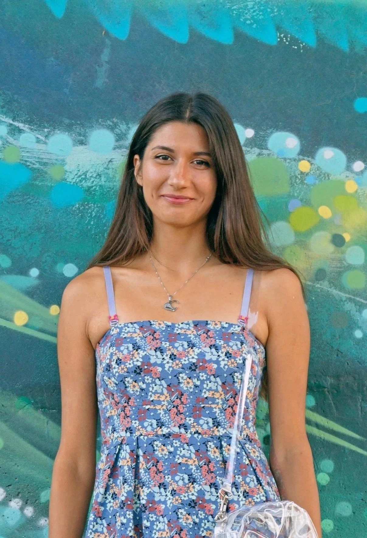 A young woman with long brown hair, wearing a sleeveless floral dress and a necklace with a bird pendant, standing in front of a colorful abstract background with blue, green, and yellow dots and shapes.