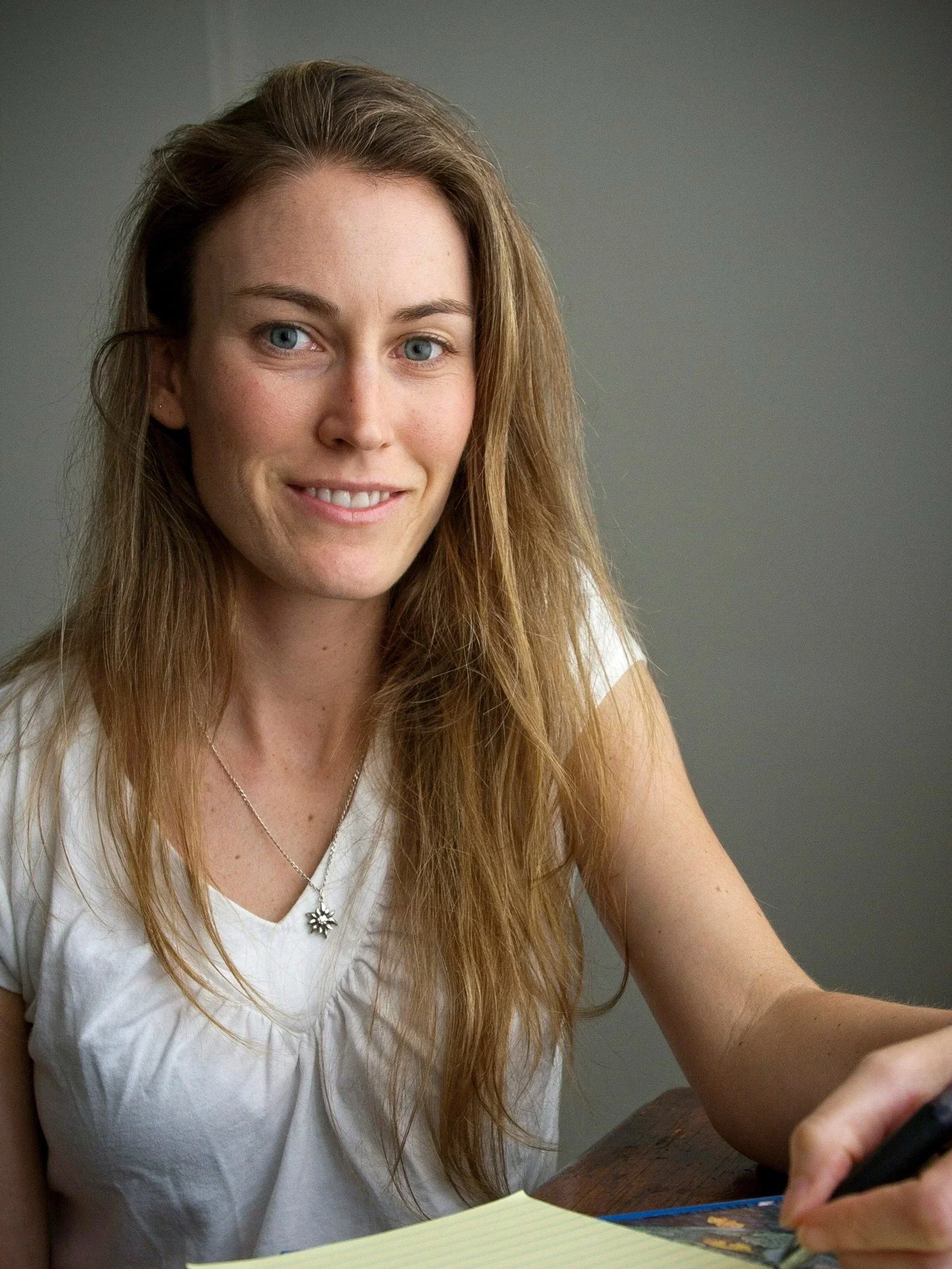A woman with long red hair, wearing a white top, smiling, and looking off to the side, sitting at a table with a yellow notebook and writing with a blue pen.