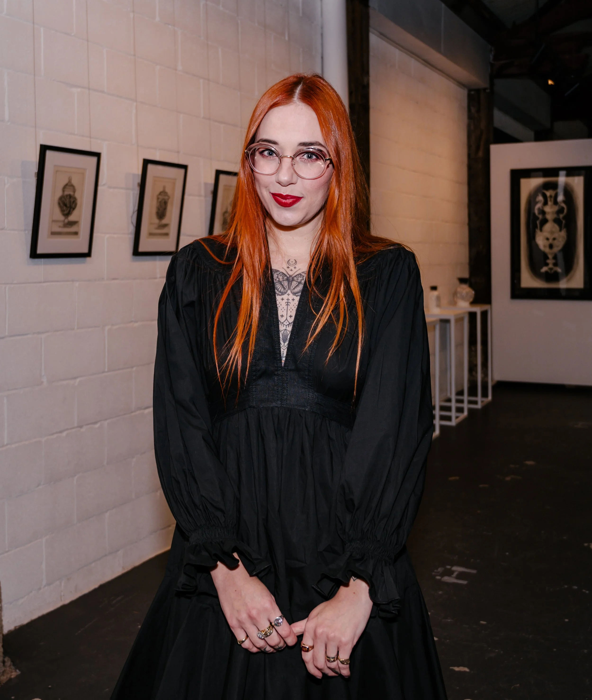 A photo of Eddy Lou with long ginger hair, black dress, glasses, and red lipstick standing in an art gallery displaying framed artworks on a white brick wall behind her. At her solo exhibition of ceramics and pen and ink drawings, in Newcastle NSW.