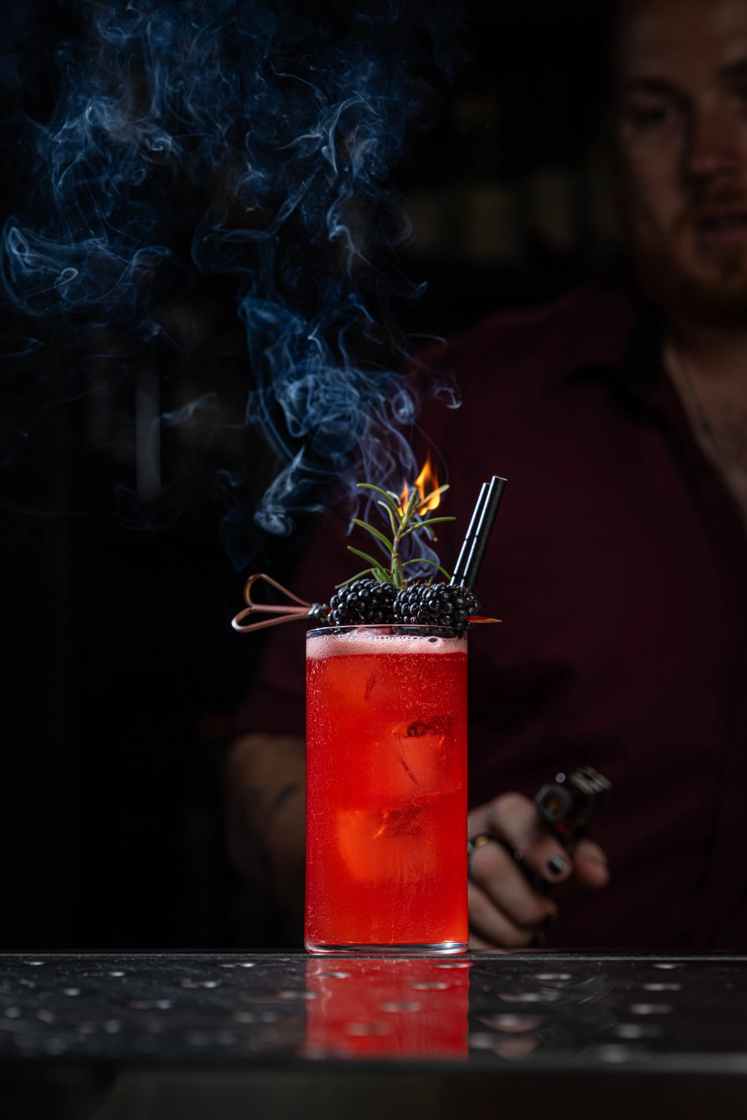 A bright red cocktail in a tall glass garnished with blackberries, a sprig of rosemary, and two black straws. Smoke rises from the top of the drink while a man in a dark background observes, holding a phone.