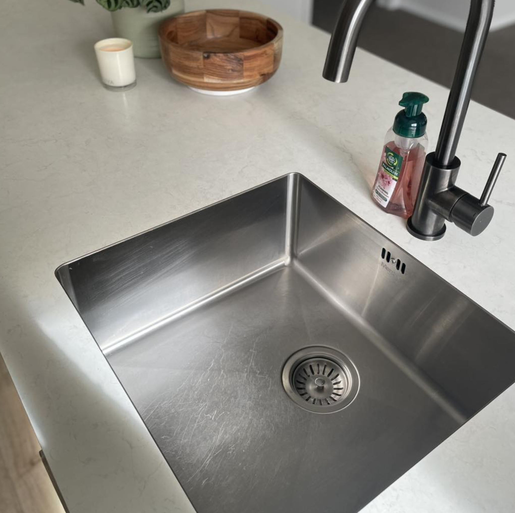 A stainless steel kitchen sink with a single basin and a modern brushed finish, installed in a white countertop with a black faucet and a pink soap dispenser. In the background, there's a small candle, a green plant, a wooden bowl, and a white container.