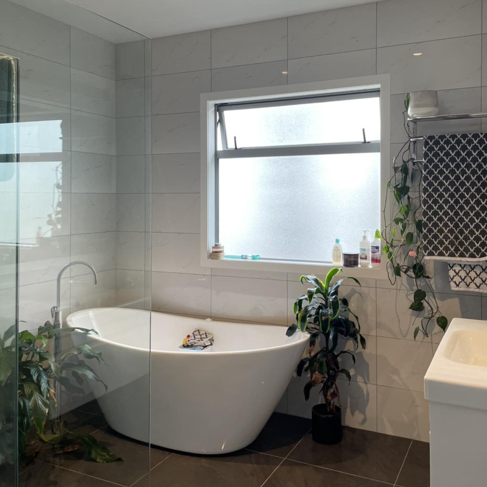 Bathroom with a freestanding white bathtub near a frosted window, plants on the floor and shelf, and a towel rack with a patterned towel.