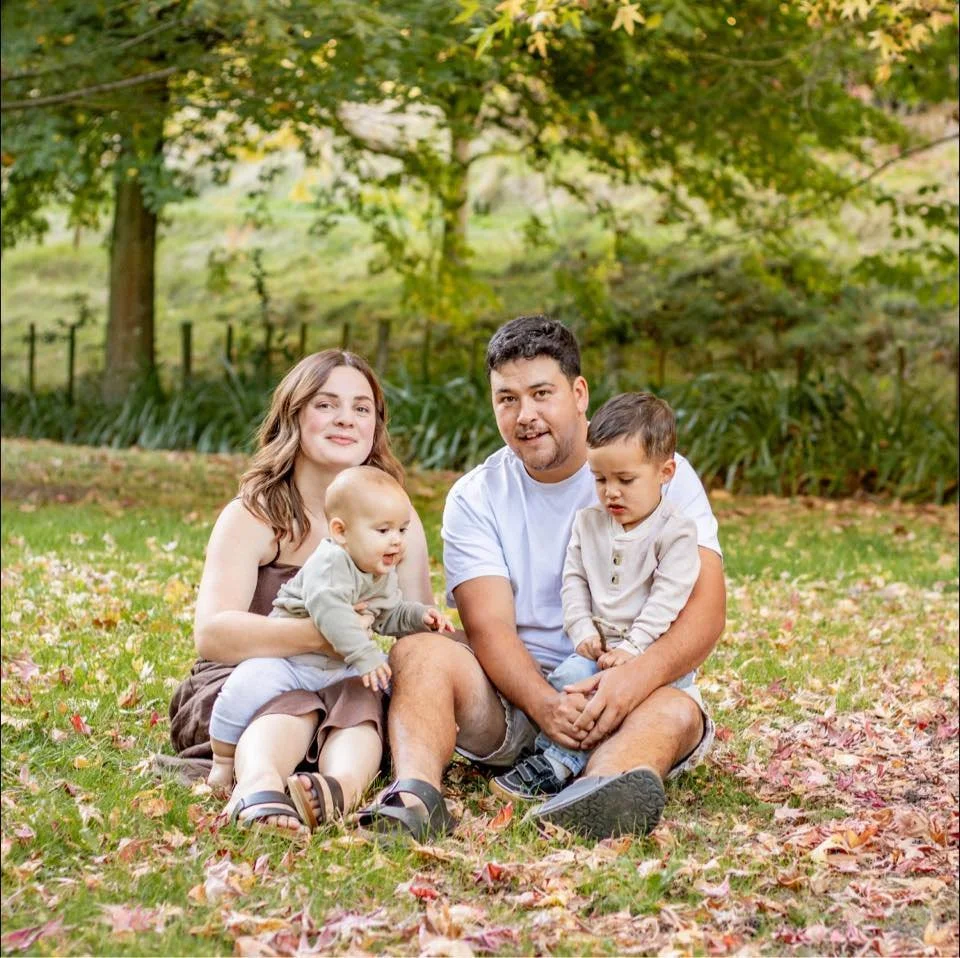 Family of four sitting on fallen autumn leaves in a park with trees in the background.