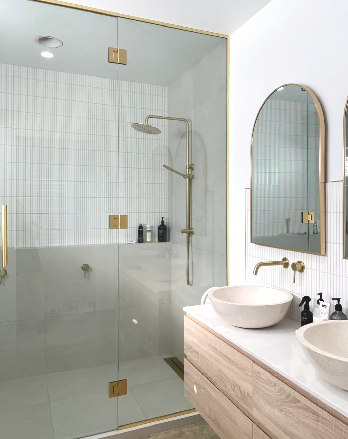 Modern bathroom with a glass-enclosed shower area, white tile walls, a wooden vanity with two round vessel sinks, and oval mirrors with gold accents.