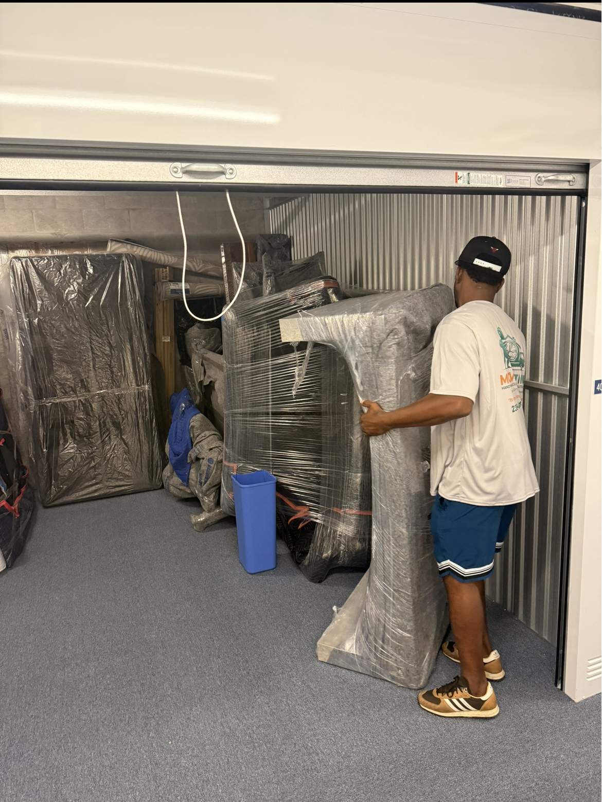 A man in a white t-shirt, blue shorts, and sneakers is stacking or handling wrapped furniture or appliances in a storage unit.