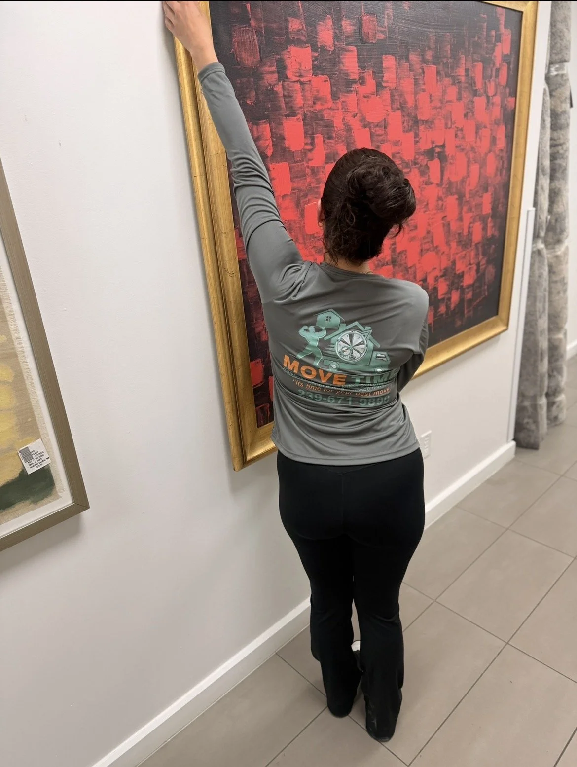 A woman in a gray long-sleeve shirt and black pants is adjusting or hanging a large abstract red and black painting with a gold frame on a white wall.