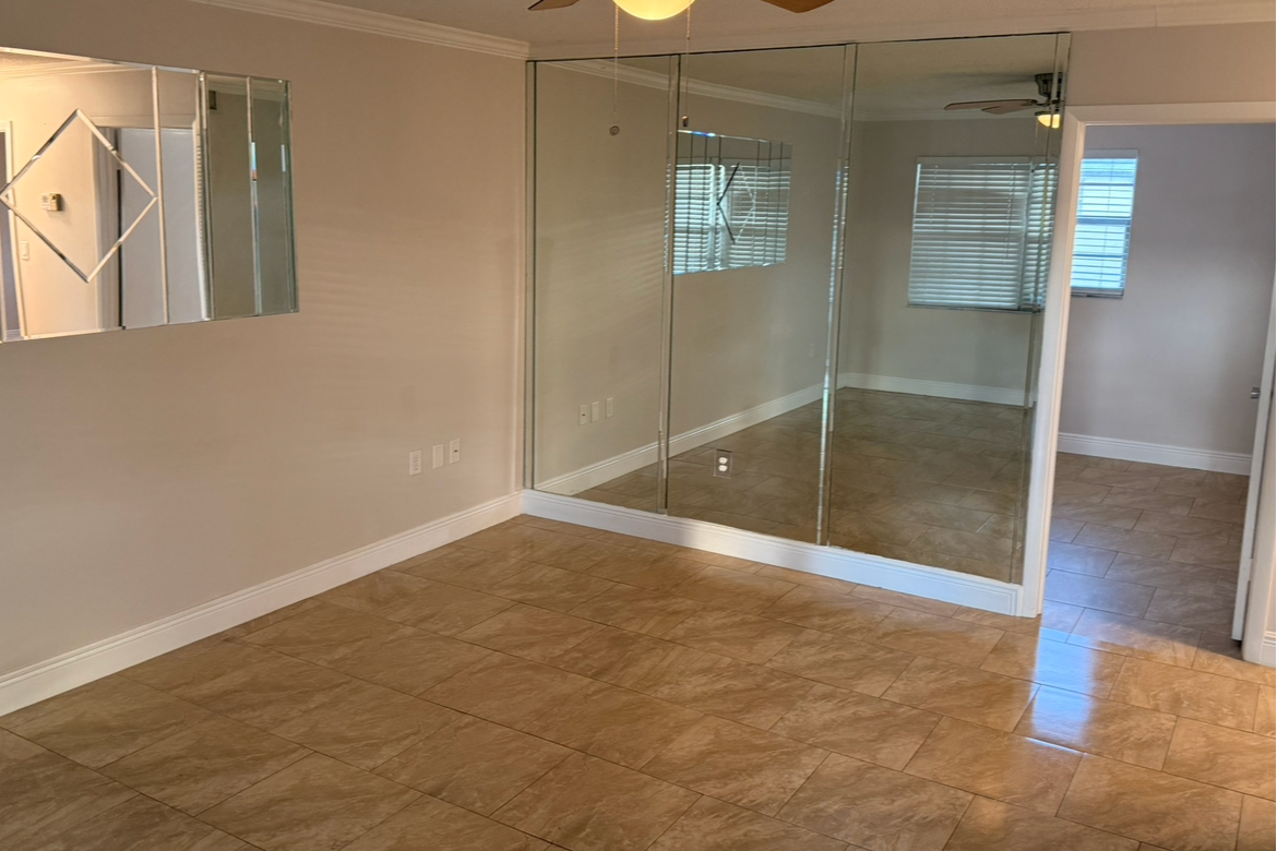 Empty room with beige walls, brown tiled flooring, a large mirror on the left wall, and glass partitions with mirrors on the right side, leading to another room with similar flooring and window blinds.