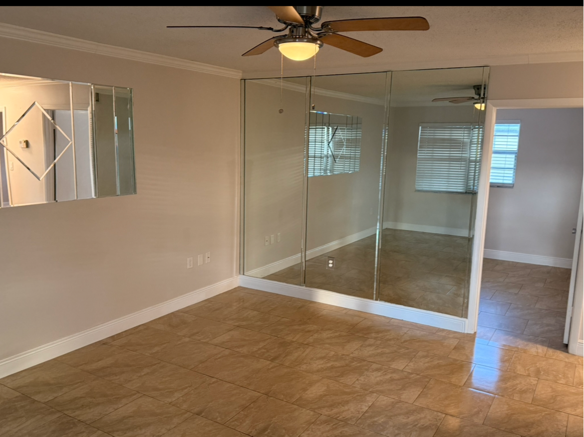 Empty living room with beige tile floors, a ceiling fan, and mirrored walls.