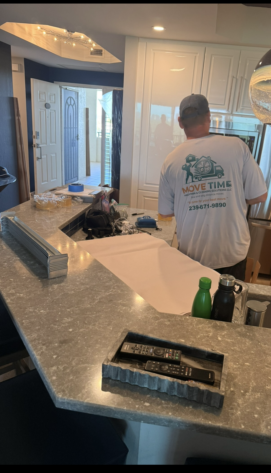 A man wearing a MOVE TIME moving company t-shirt standing in a kitchen, seen from the back, with various items on a gray marble countertop, including remote controls in a tray, water bottles, and miscellaneous objects. The kitchen has white cabinets and an open door leading outdoors.