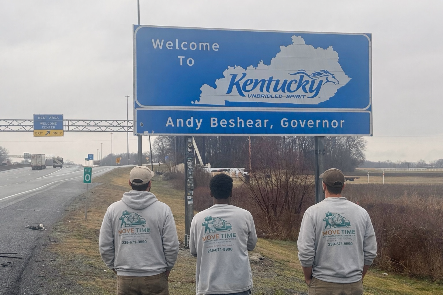 Three people standing near a highway sign that says 'Welcome to Kentucky,' with a map outline and the words 'Unbridled Spirit,' and the name 'Andy Beshear, Governor.'