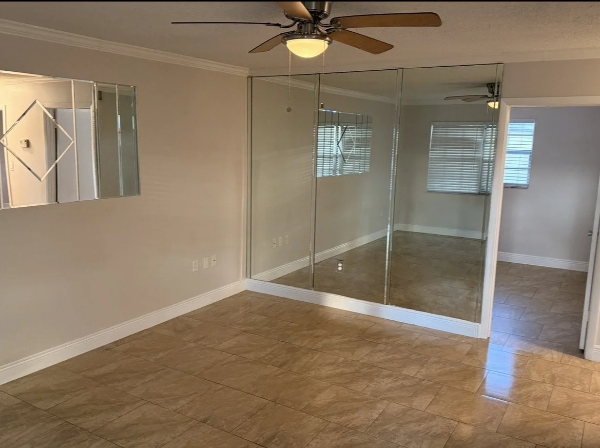 Empty living room with beige walls, wooden floor tiles, a ceiling fan, and a mirrored closet door.
