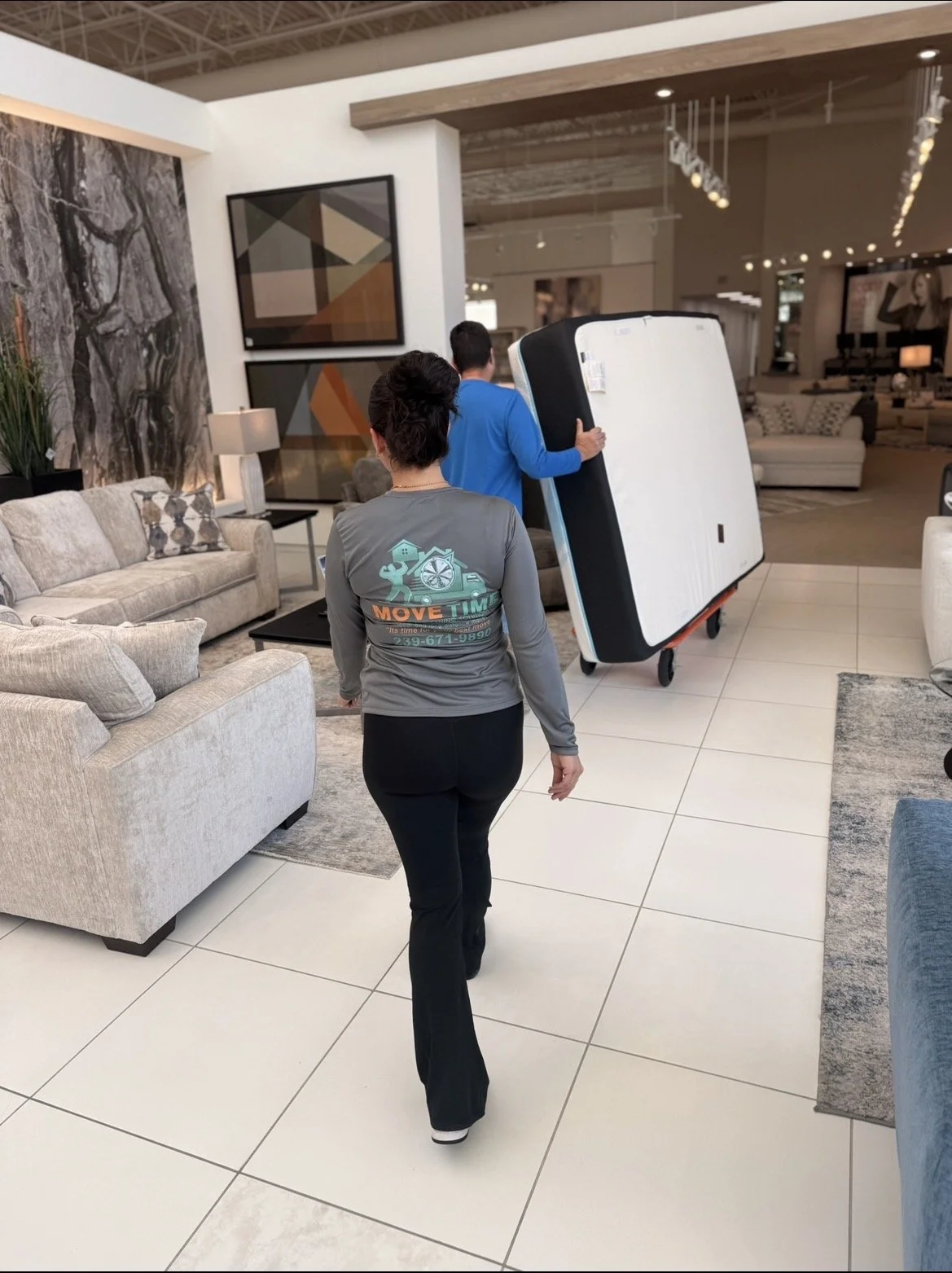 Two people carrying office furniture, possibly beds, through a furniture store with living room furniture and artwork in the background.