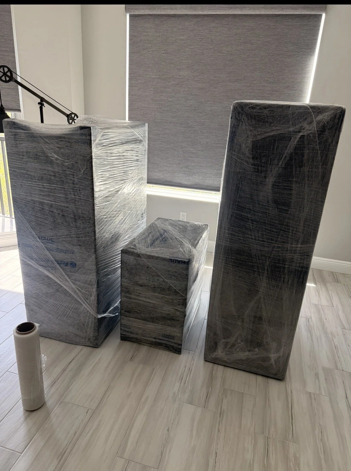 Four wrapped pieces of furniture, including two tall rectangular cabinets, one shorter cabinet, and a small box, placed on a light-colored wood floor near a window with a closed gray blind.