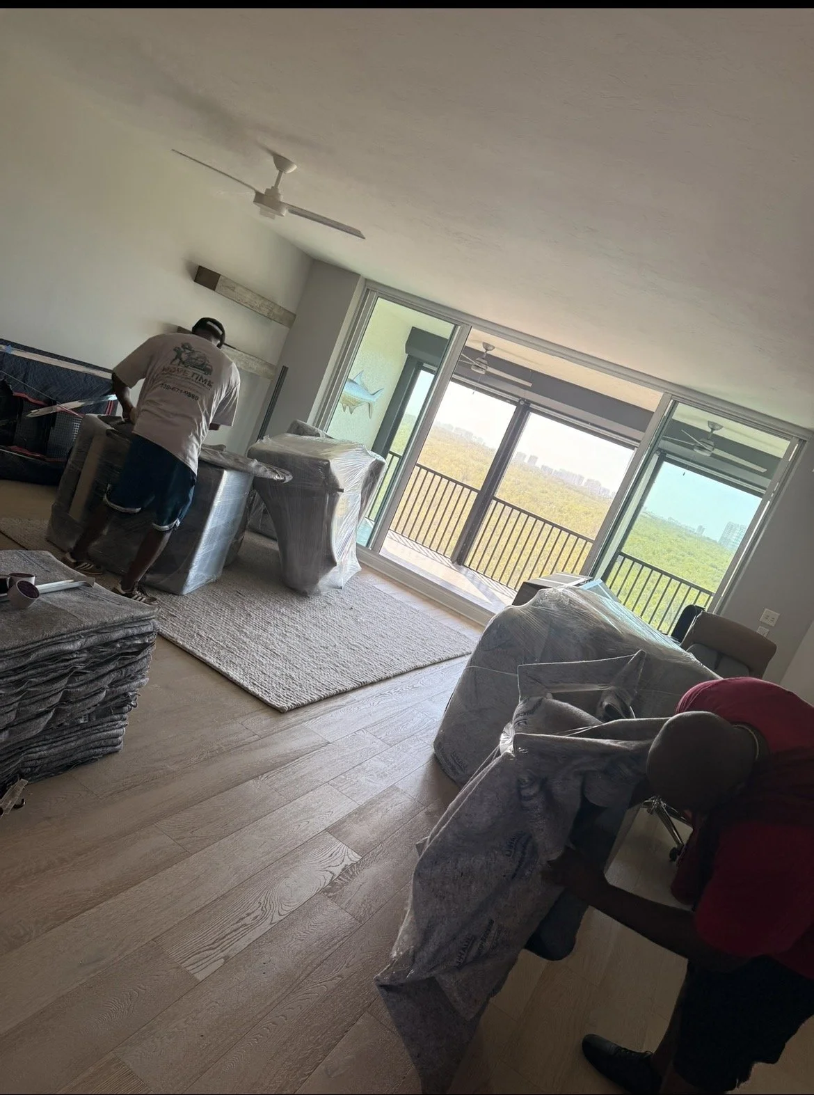 Two workers in a living room with large sliding glass doors, some furniture wrapped in plastic, and worker stacking or arranging items.