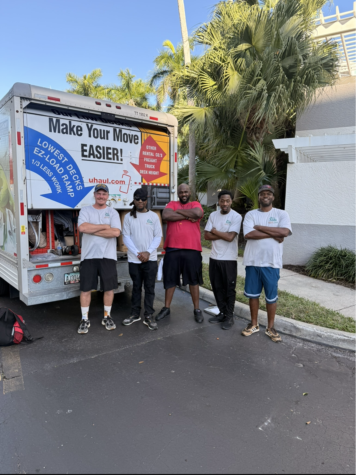 Five men standing in front of a U-Haul truck, smiling, with palm trees and a white building in the background. The truck has signs promoting moving services.