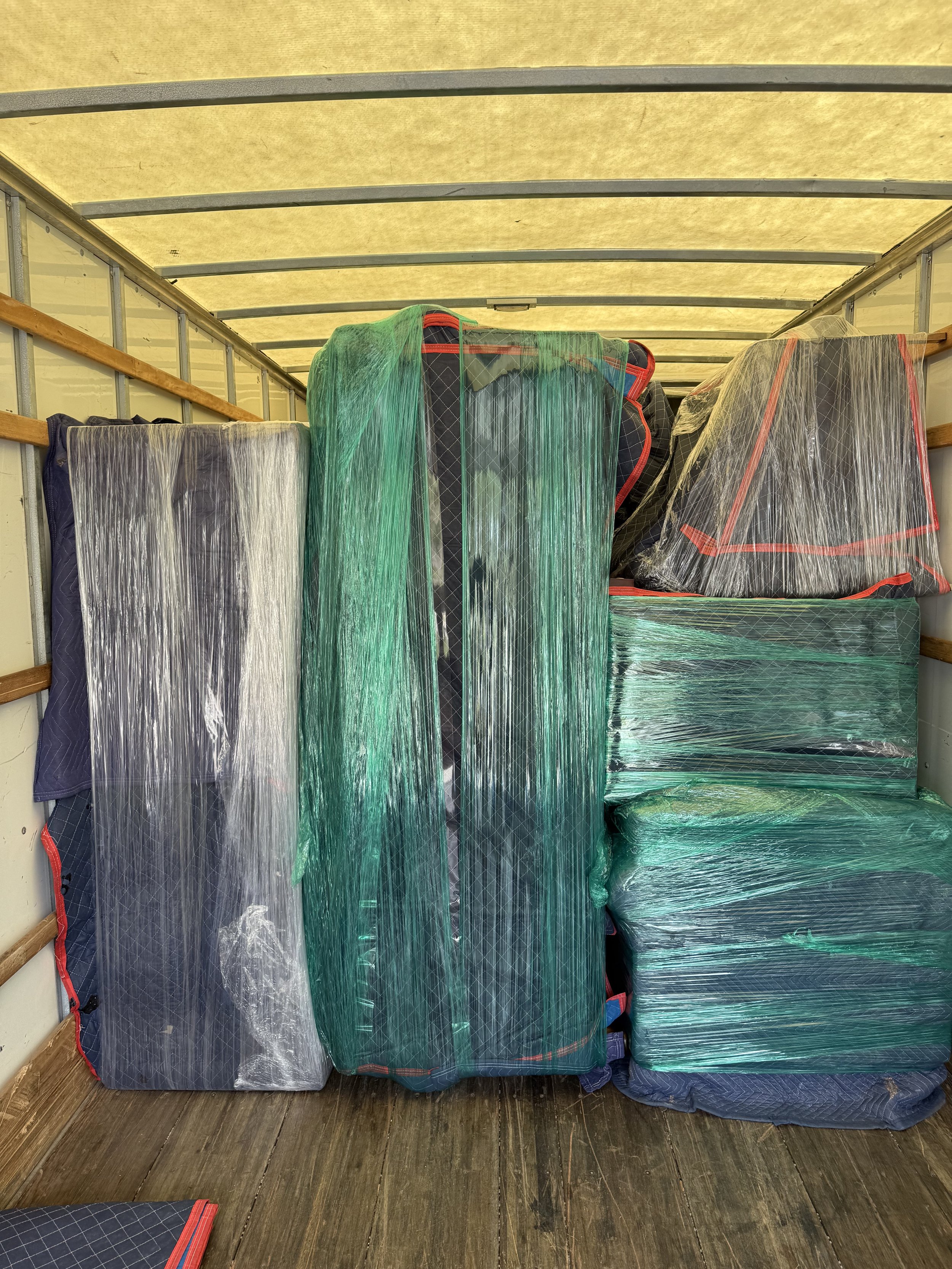 Several suitcases and bags wrapped in green and clear plastic, stored inside a moving truck with wooden side rails and a wood floor.