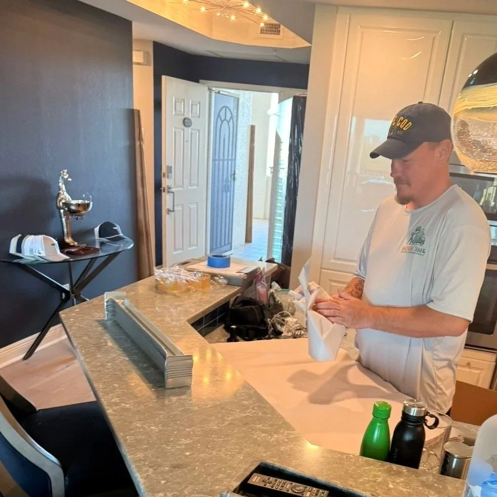A man wearing a gray t-shirt and cap standing at a kitchen counter, folding a napkin. The kitchen includes a marble countertop, white cabinets, and a decorative hanging light. In the background, there is a door leading outside and a small table with a trophy and hats.