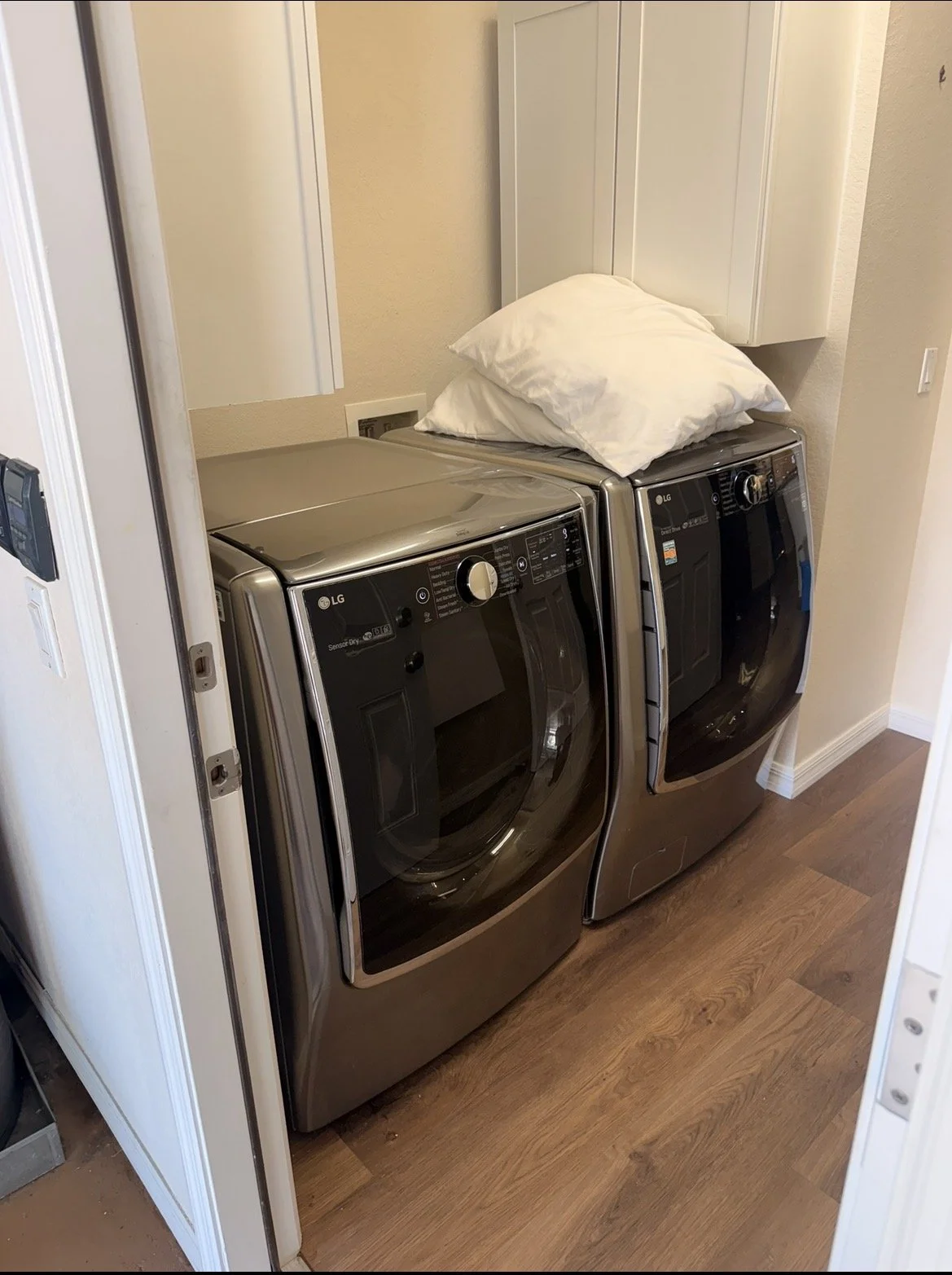 A laundry room with two stainless steel LG washing machines, one stacked on top of the other. There are two white pillows placed on top of the machines. The room has a beige wall and wooden flooring.