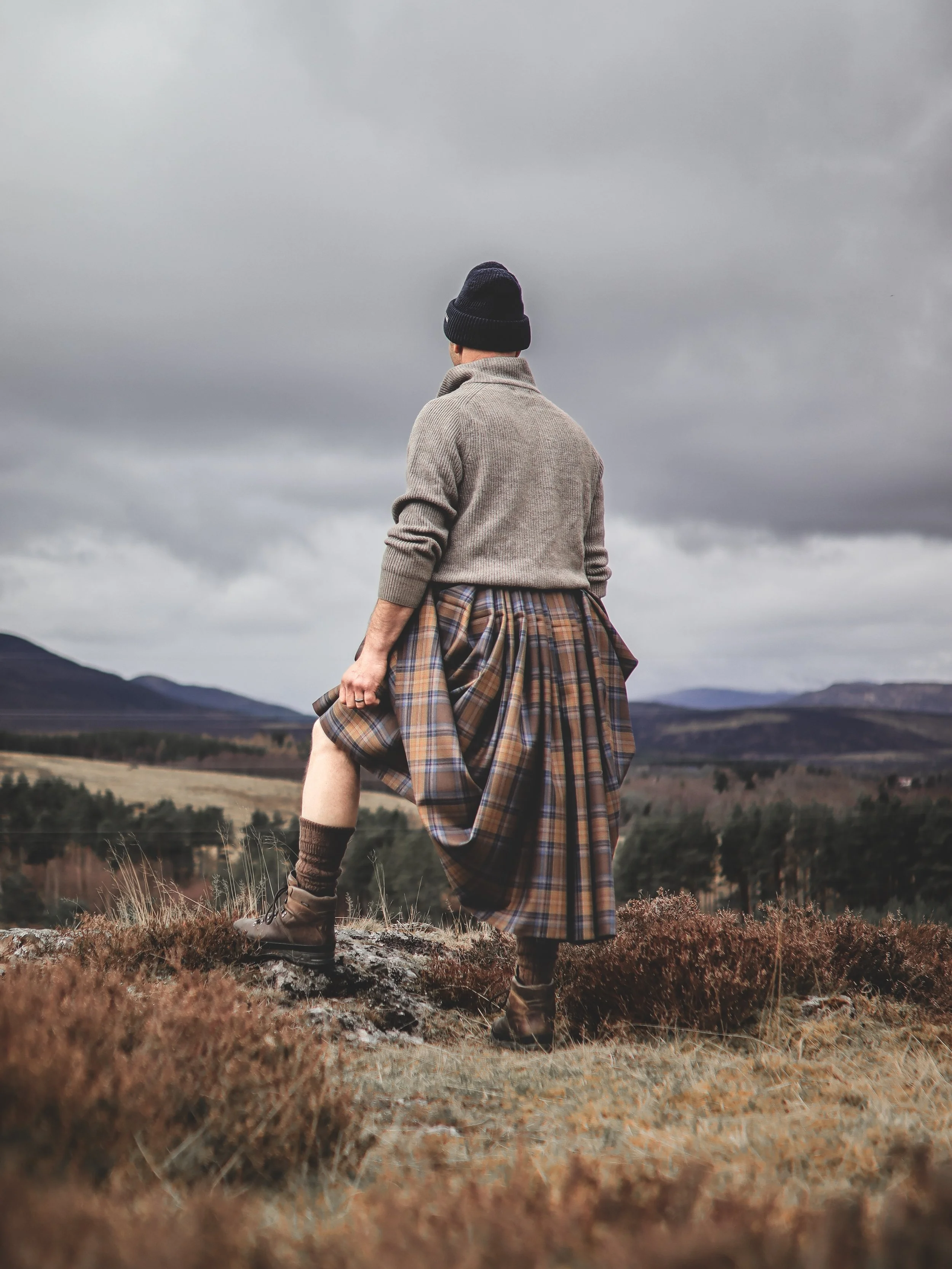 Outdoor portrait photography of a person wearing a tartan kilt standing on a hillside in the Scottish Highlands, overlooking a dramatic mountain landscape.