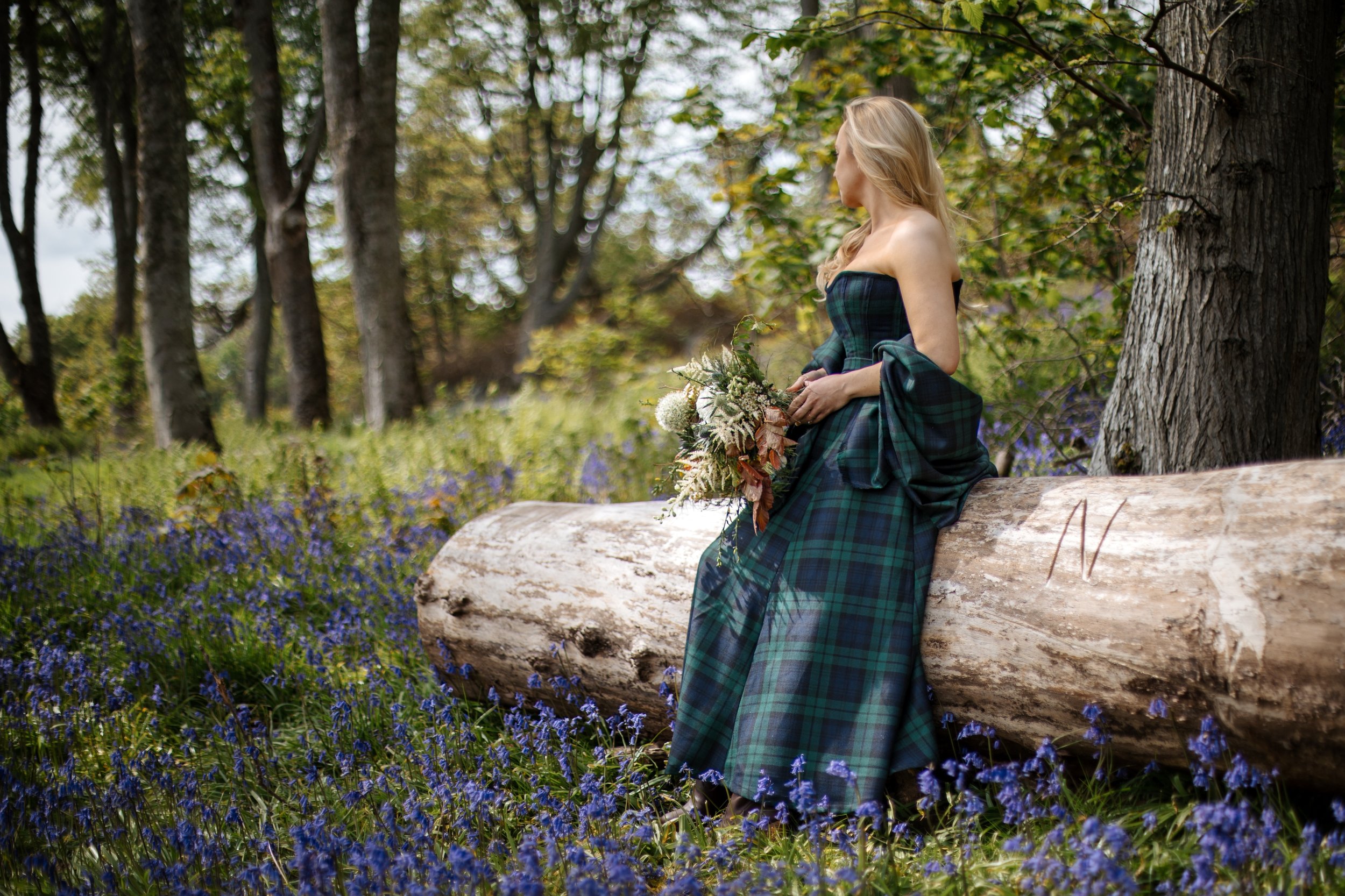 Outdoor portrait photography of a woman wearing a Thistle and Cloth tartan dress seated on a fallen tree among bluebells in a Scottish woodland, holding a bouquet of seasonal flowers.