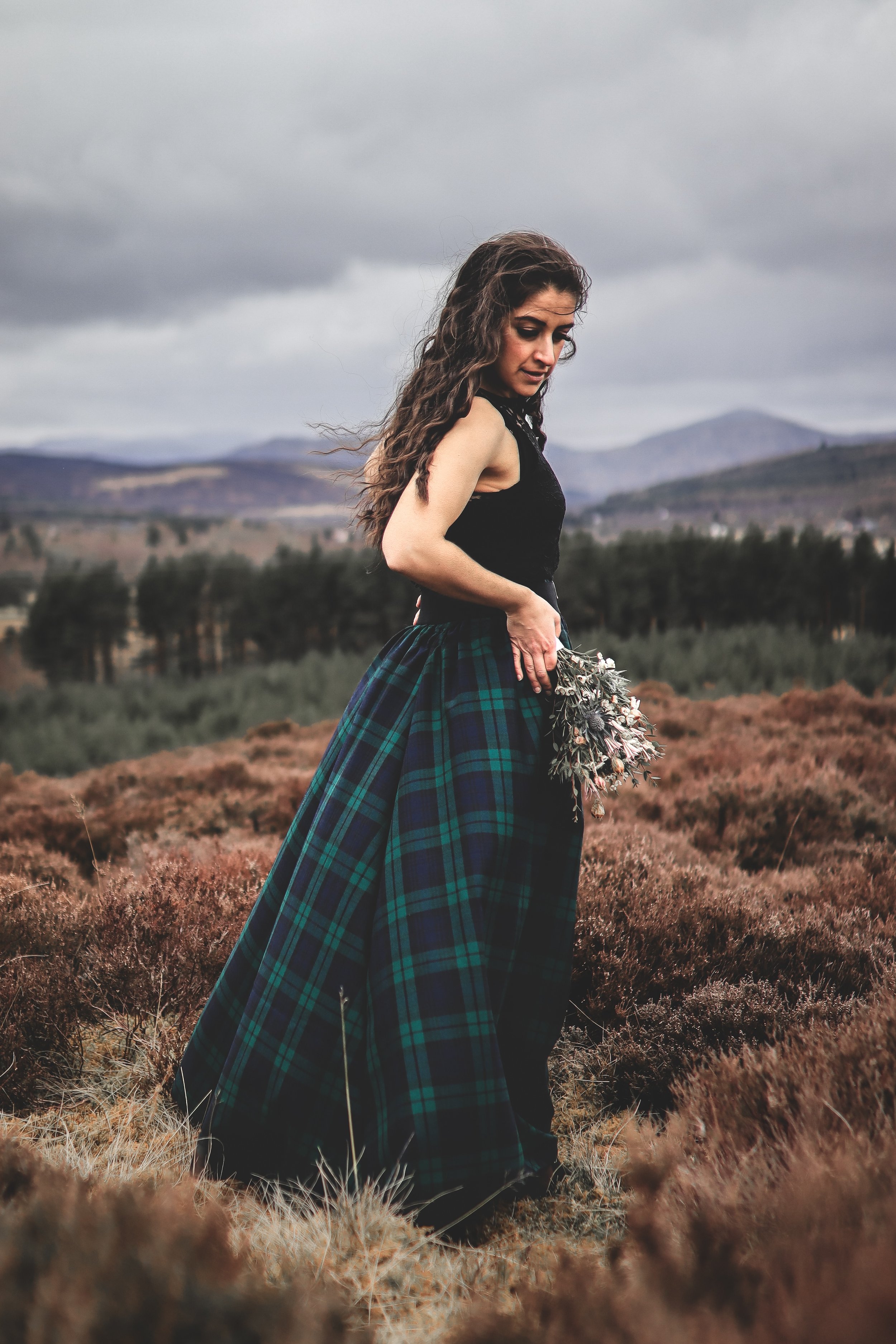 Outdoor portrait photography of a woman wearing a Thistle and Cloth tartan skirt standing in the Scottish Highlands, surrounded by heather with mountains in the background.