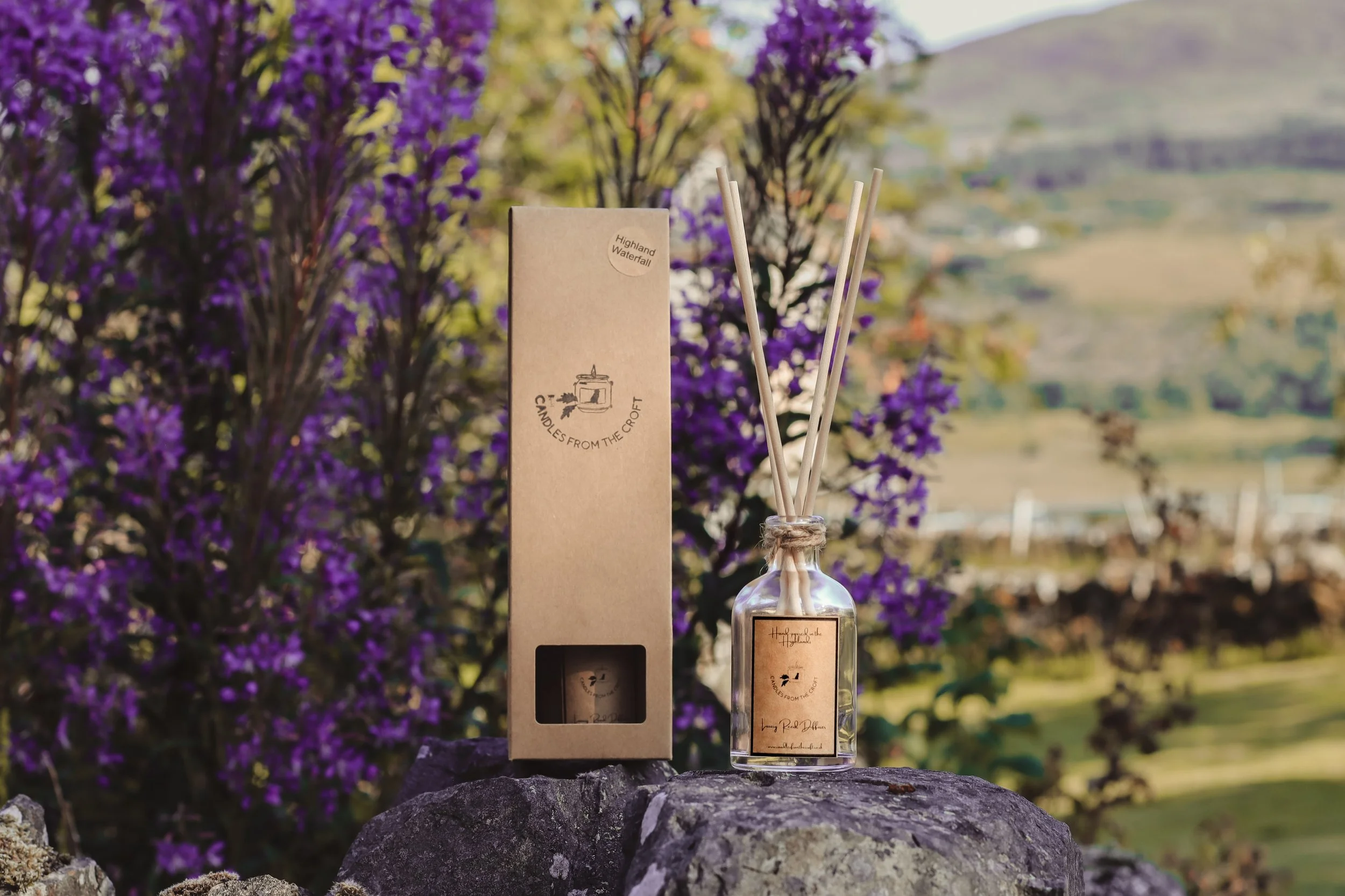 Product photography of a Candles from the Croft reed diffuser displayed on a stone wall with vibrant purple flowers and a Highland landscape in the background.