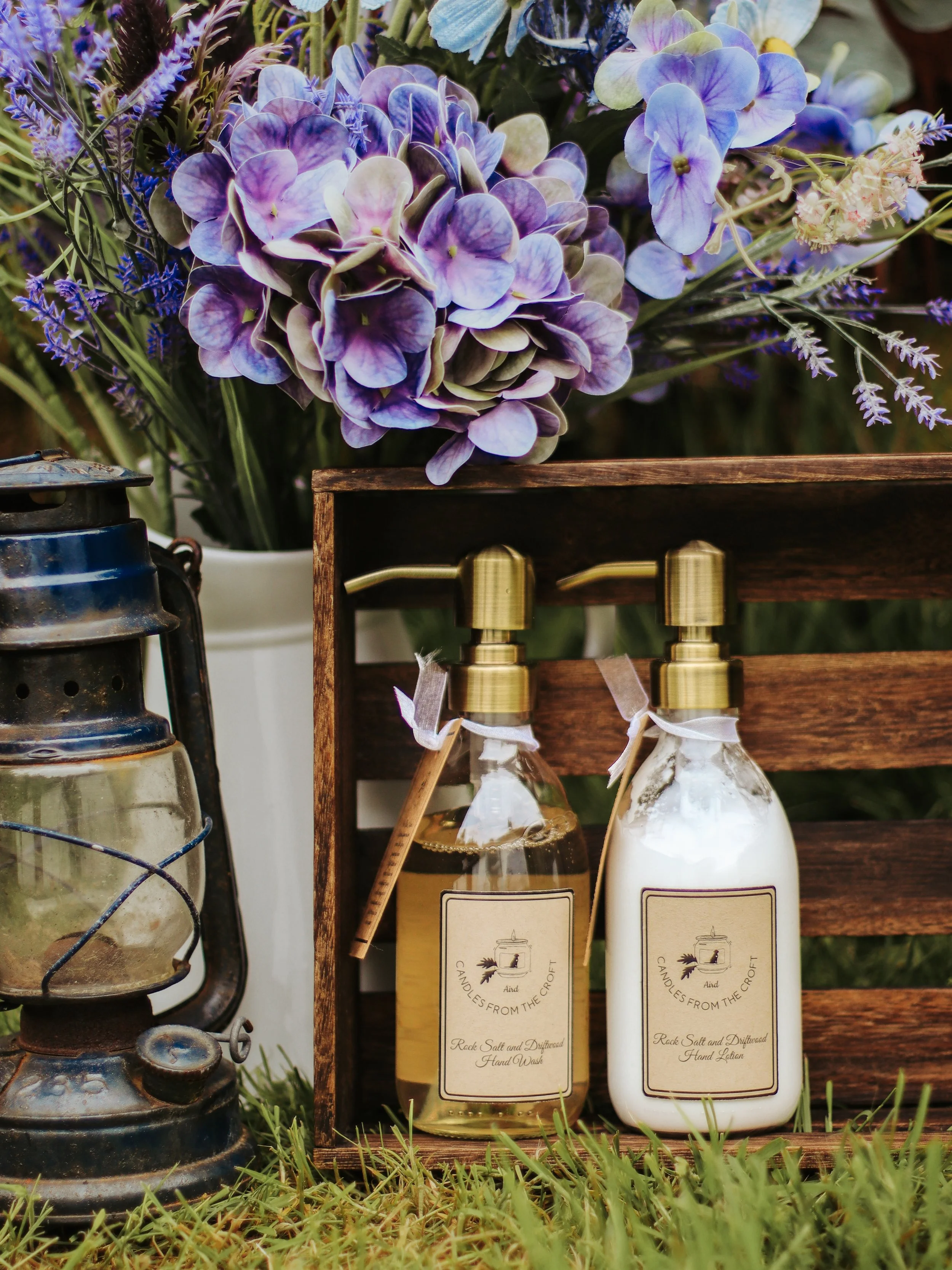 Product photography of Candles from the Croft hand wash and hand lotion displayed in a wooden crate with purple flowers and a vintage lantern.