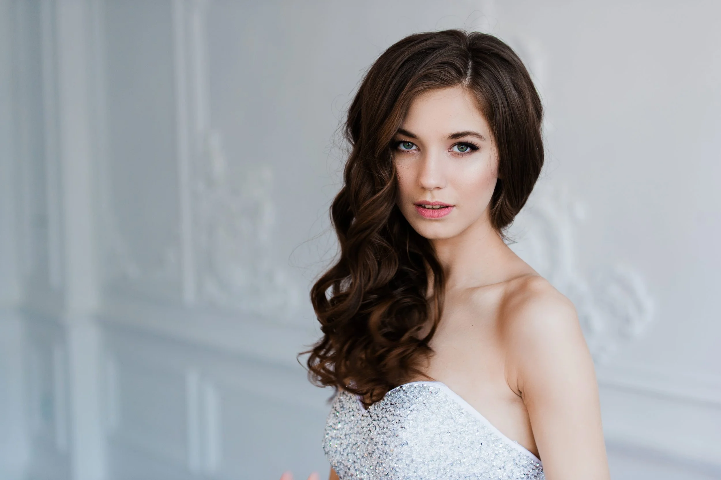 A young woman with long, wavy brown hair and blue eyes looking at the camera, wearing an off-shoulder silver sequined dress, standing in front of a plain light-colored background.