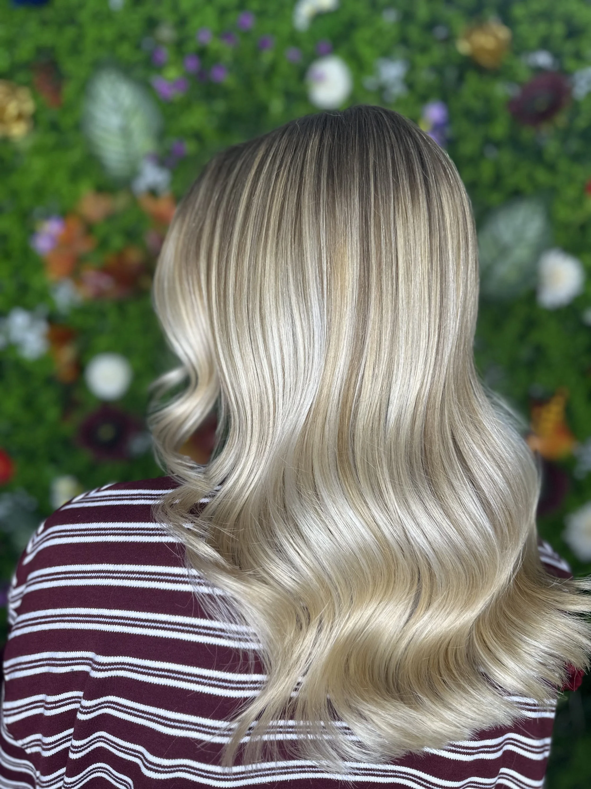 Back of a person with long, wavy, blonde hair, wearing a striped maroon and white top, standing in front of a green floral background.