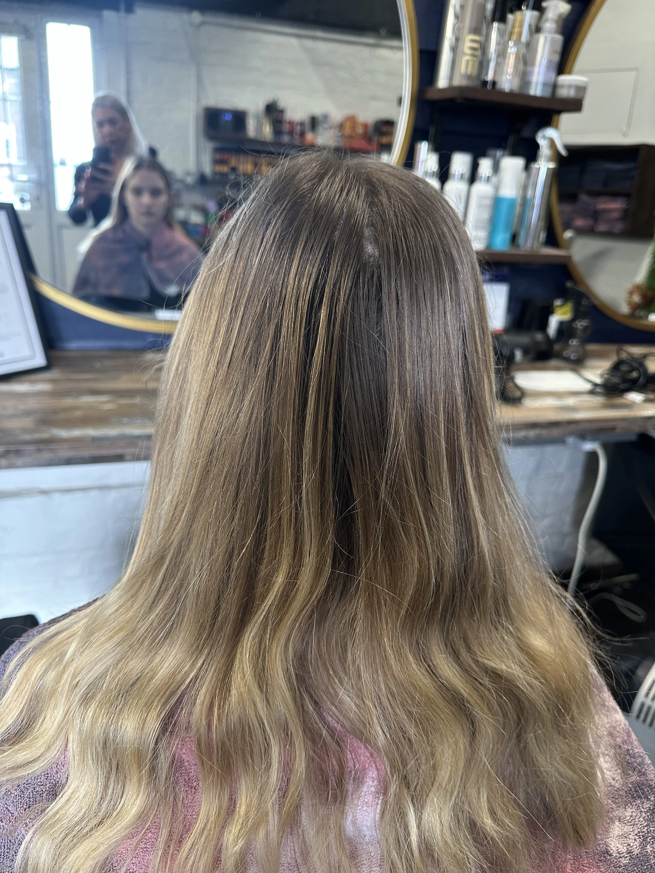 Front view of a woman's head showing long, wavy blonde hair with darker roots in a hair salon, with salon tools and products visible in the background.