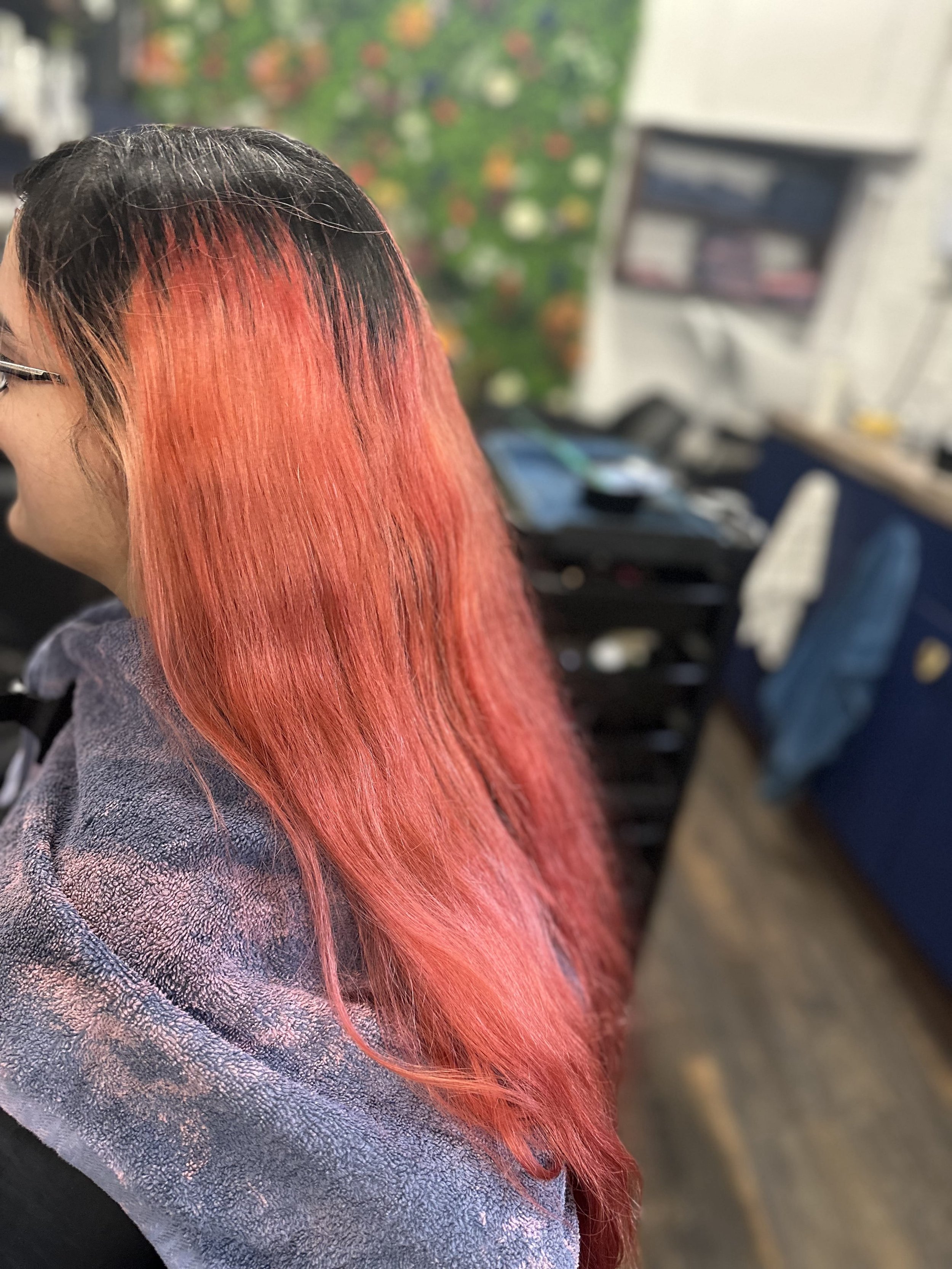 Close-up of a person's head with pink hair, wearing glasses, sitting in a salon or hair studio.