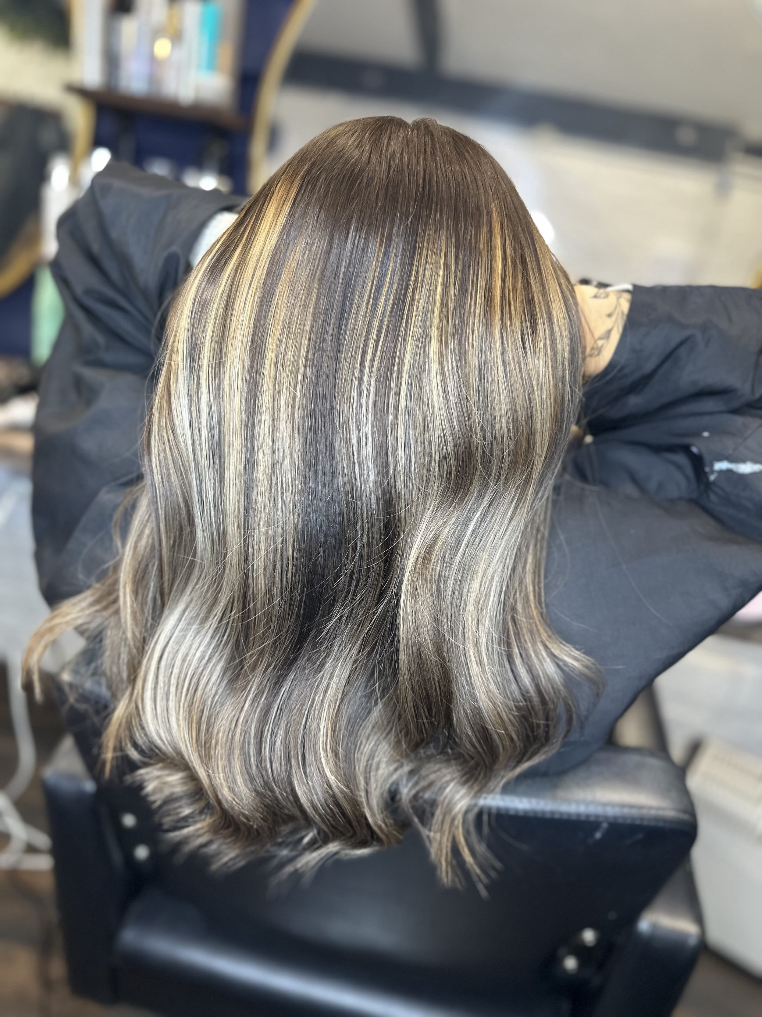 View of a person's head with long, wavy hair that is a mix of black, blonde, and gray, styled with soft waves, in a salon setting.
