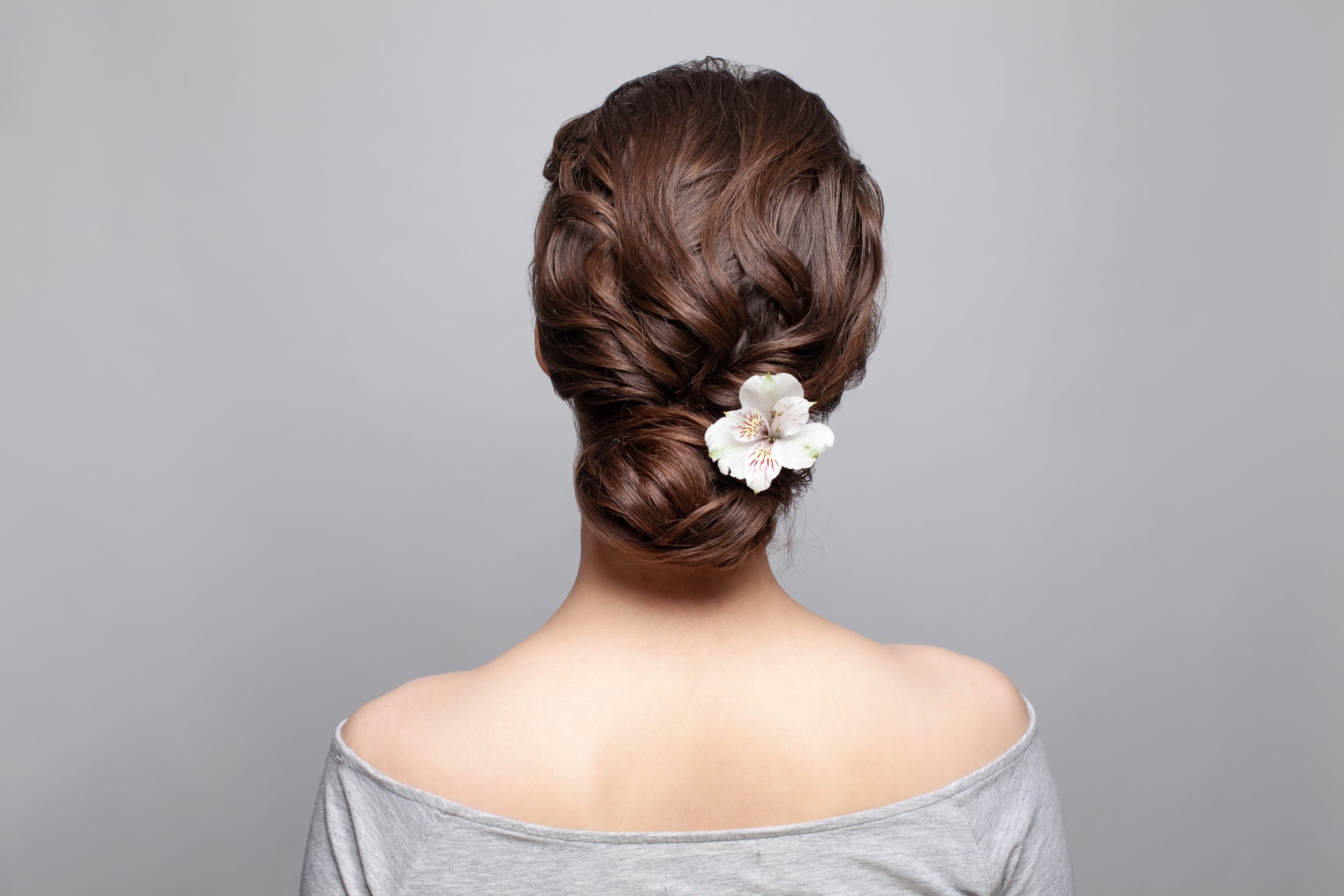 Back of a woman's head with styled brown hair, decorated with white flowers, against a plain gray background.