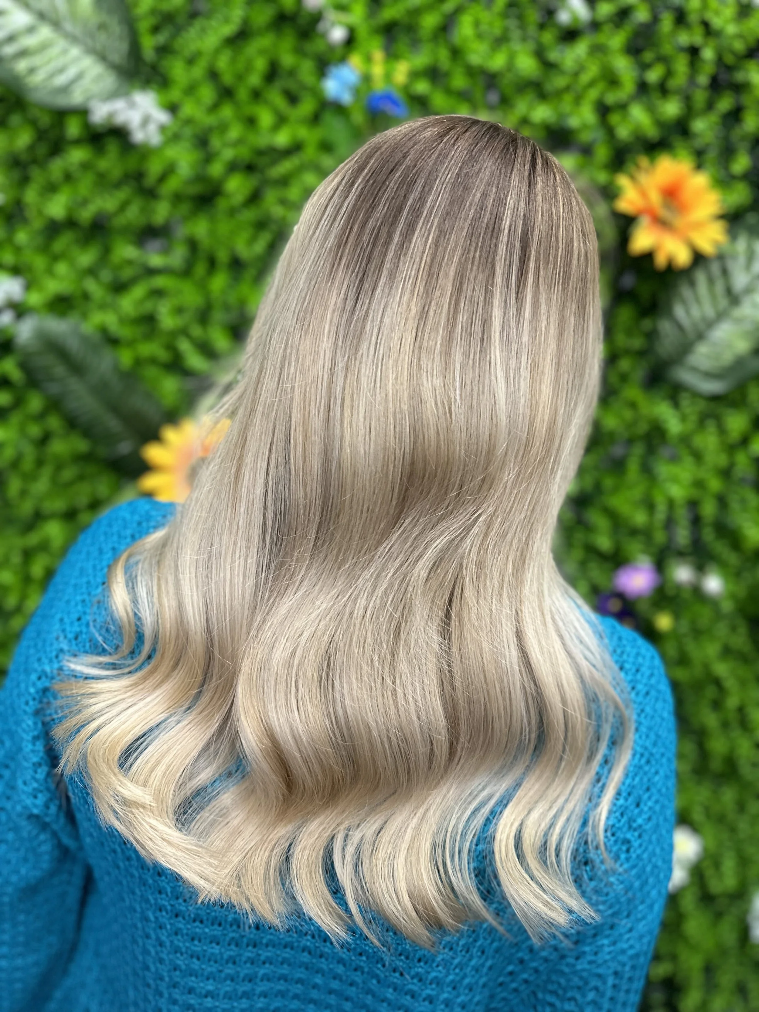 Back view of a woman with long, wavy blonde hair wearing a blue sweater, standing in front of green foliage with colorful flowers.