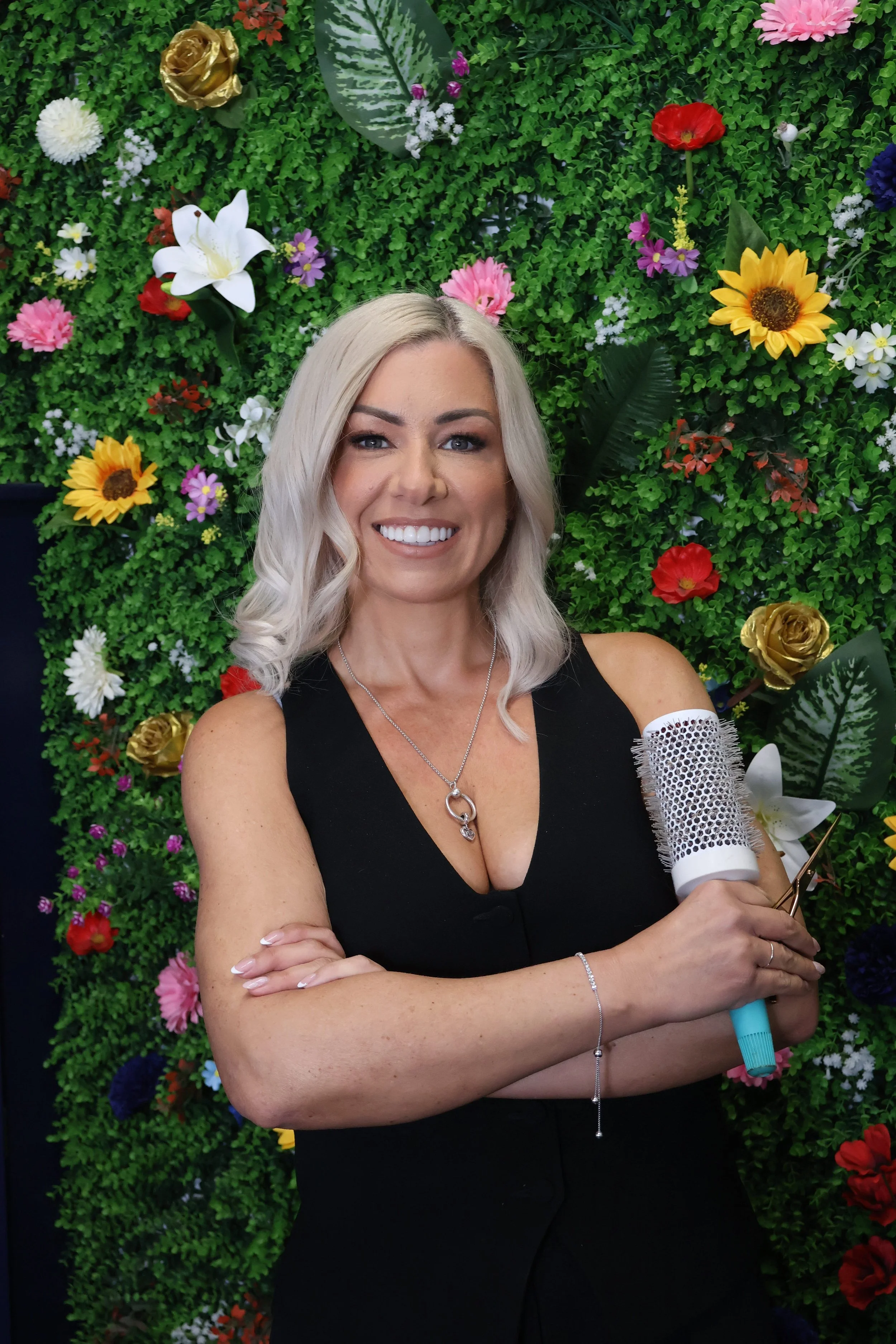 A woman with long blonde hair smiling, standing in front of a green floral wall with colorful flowers, holding a hairbrush.