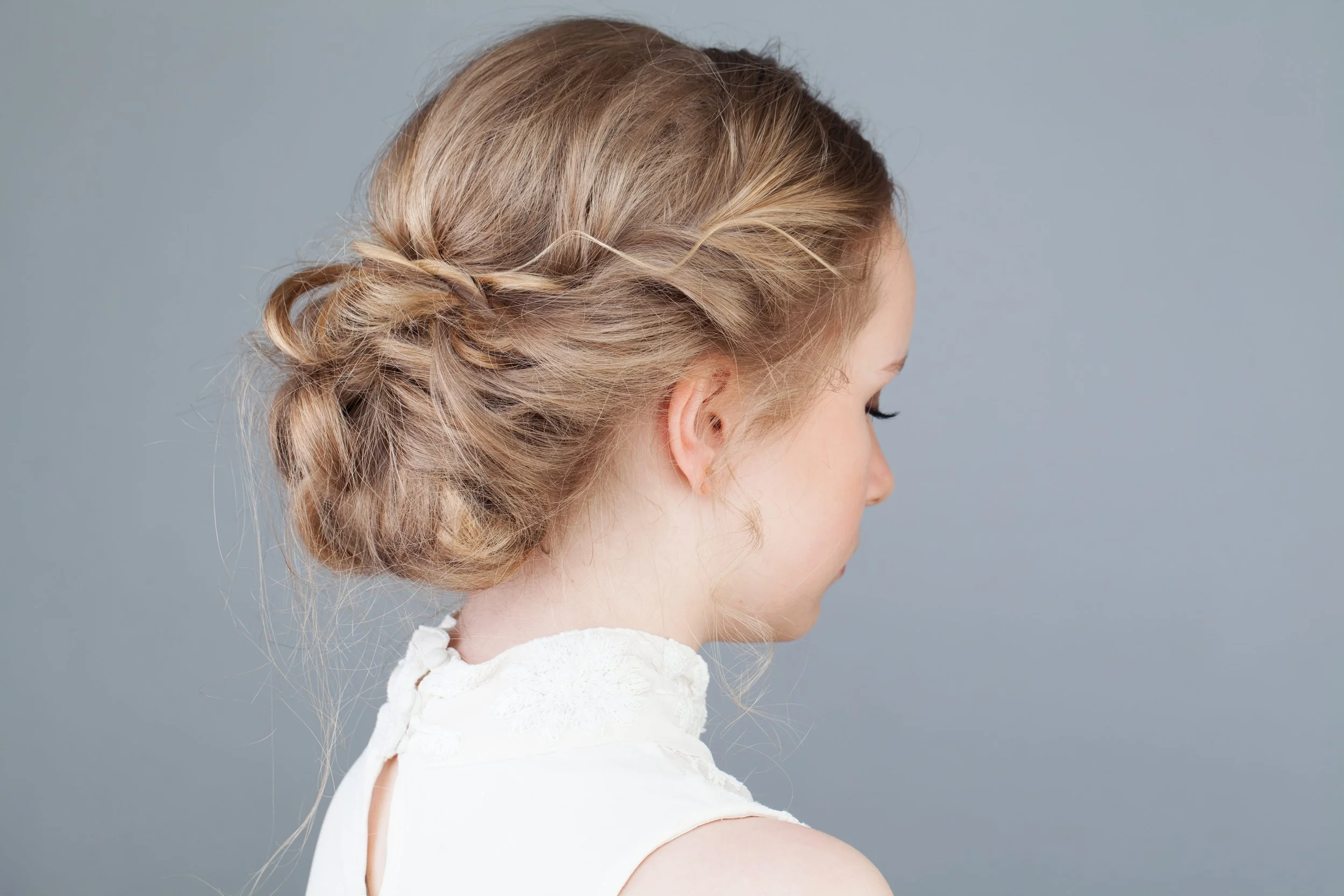 A young girl with blonde hair styled in a loose updo with soft waves, wearing a white dress with a textured shoulder detail, against a plain light gray background.