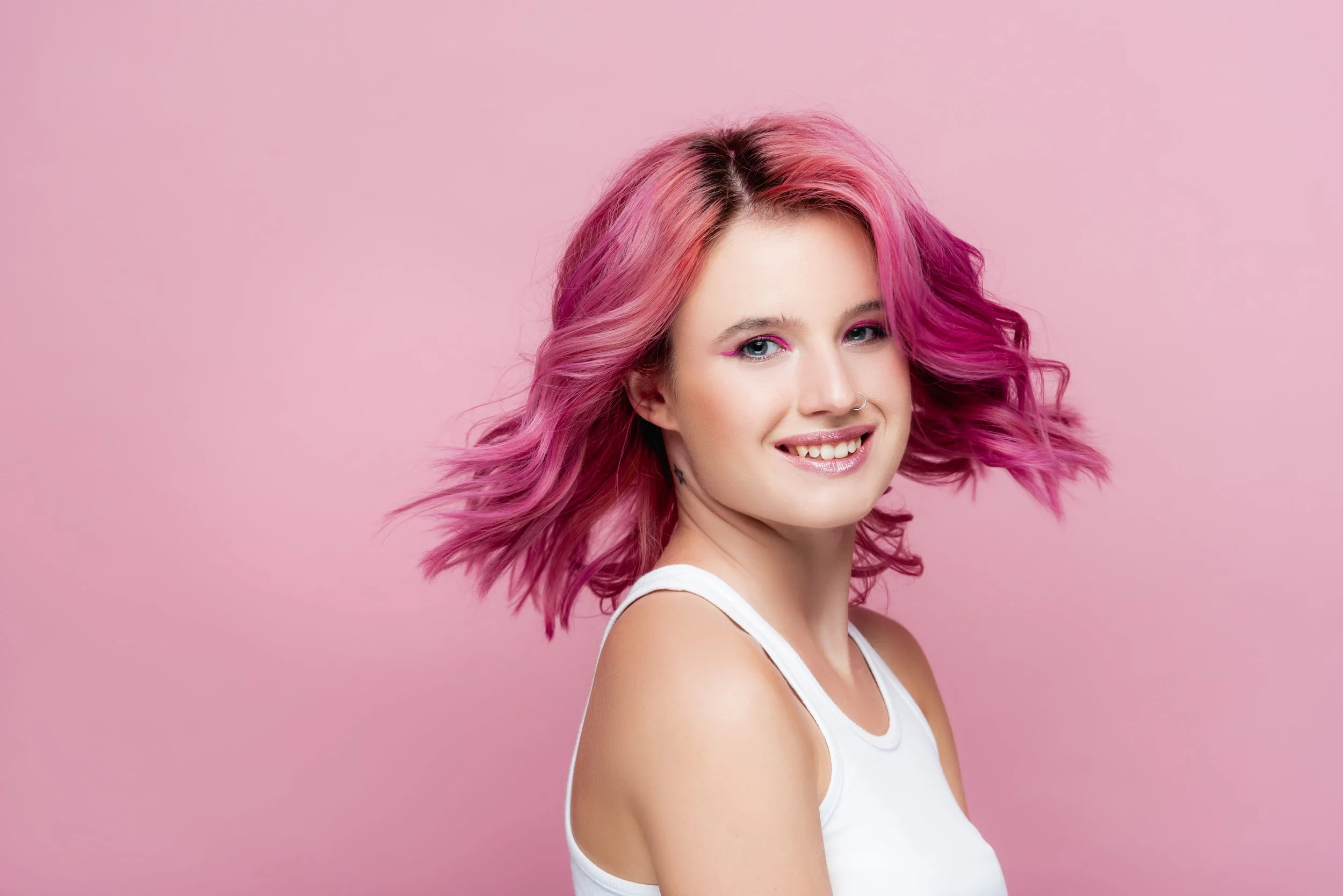 A woman with wavy pink hair smiling against a pink background.