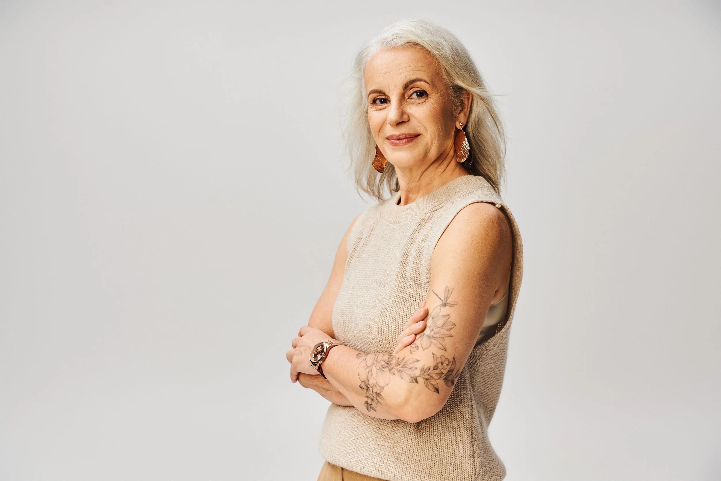 An older woman with long gray hair, wearing a sleeveless beige sweater and large earrings, standing against a plain light gray background, smiling with arms crossed and a floral tattoo on her left arm.