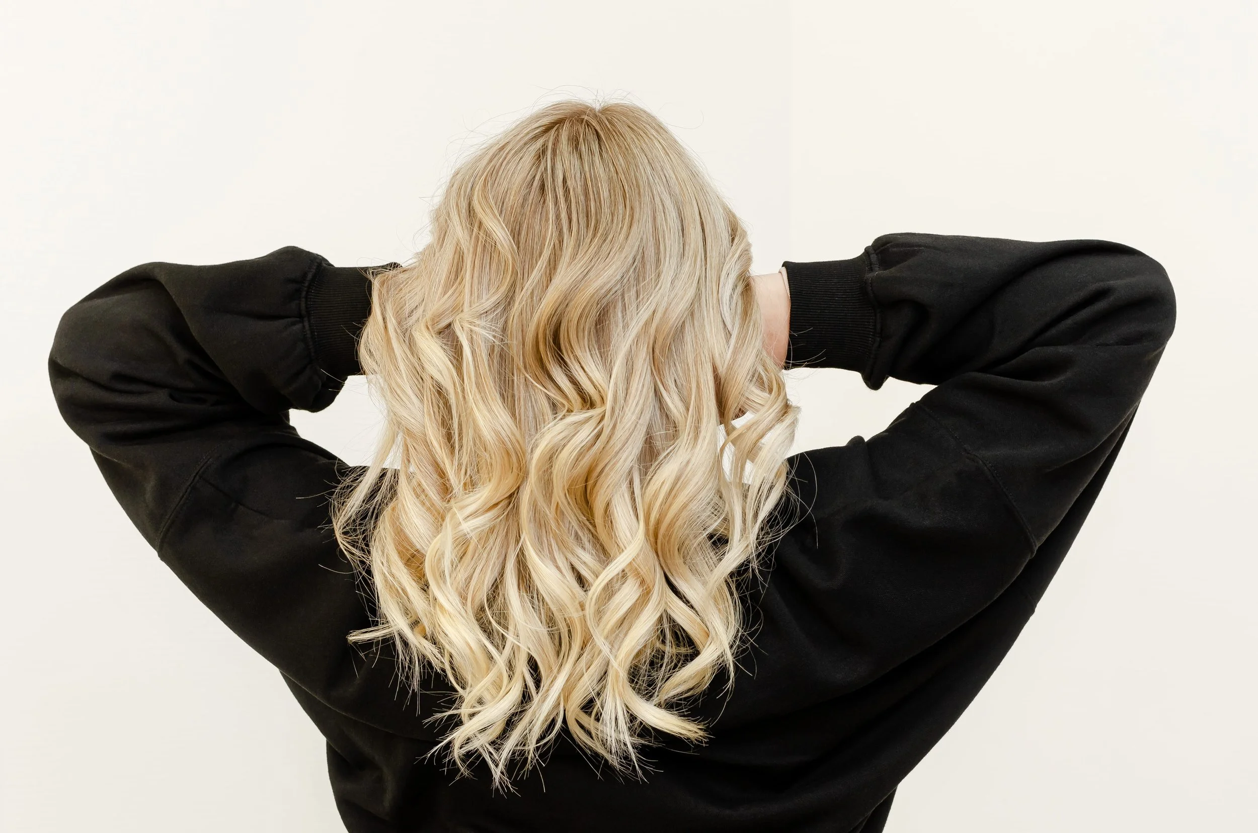 A person with long, blonde, curly hair wearing a black sweatshirt, facing away with hands behind their head, against a plain off-white background.