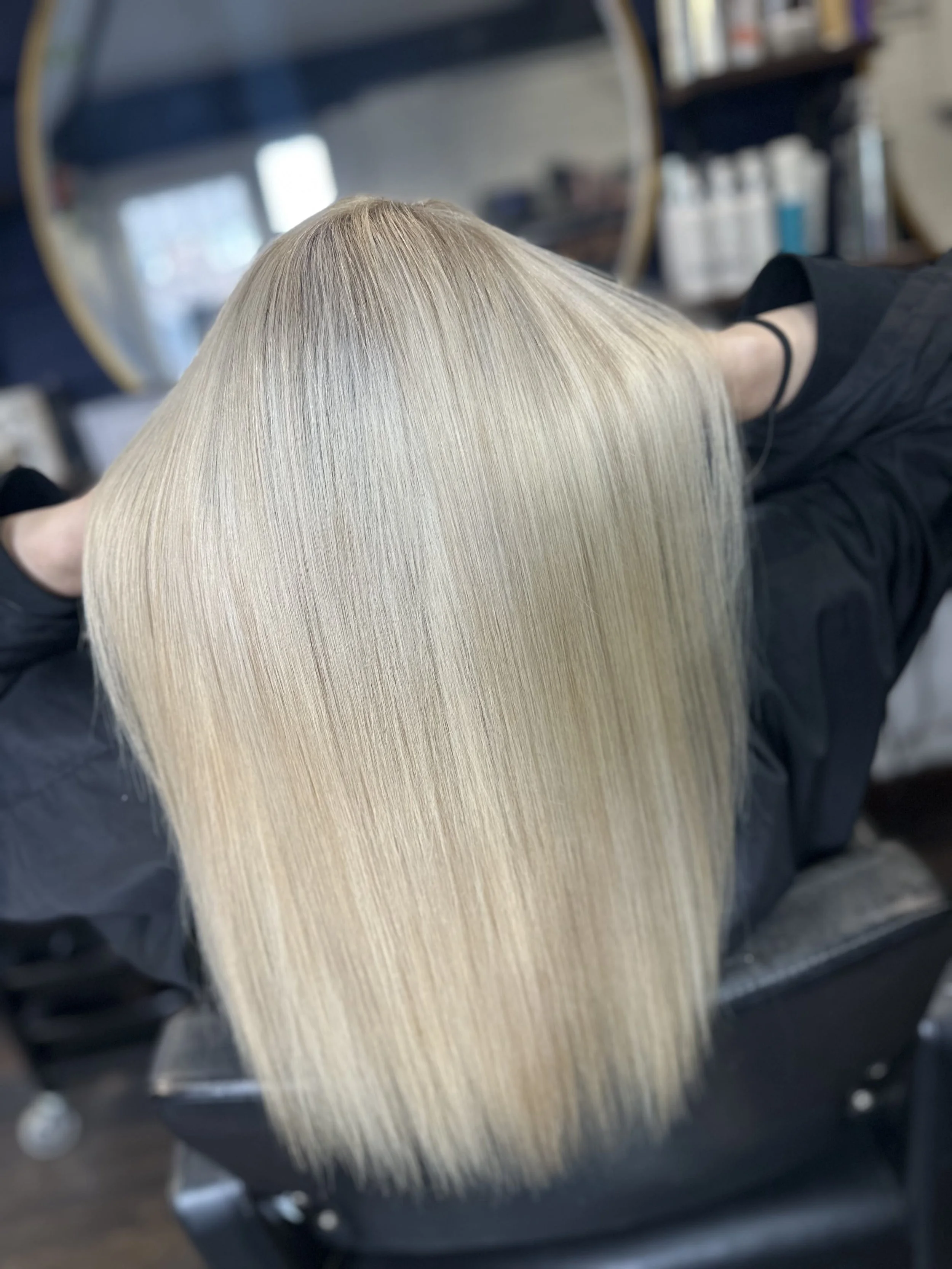 Close-up of a person's long, straight blonde hair with a mirror and salon products in the background.