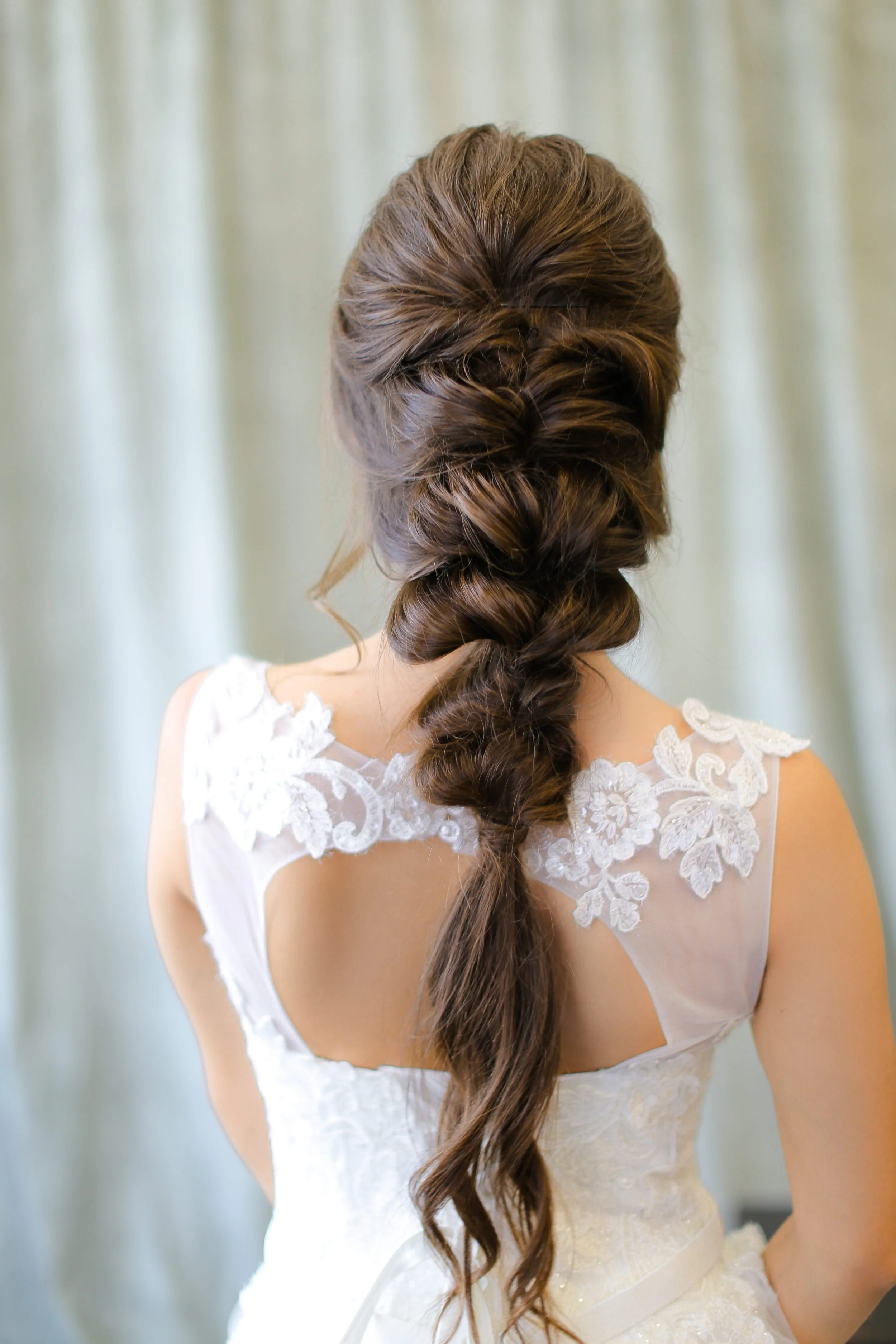Back view of a woman with long, wavy brown hair styled in a loose braid, wearing a white lace wedding gown with floral appliqué details.