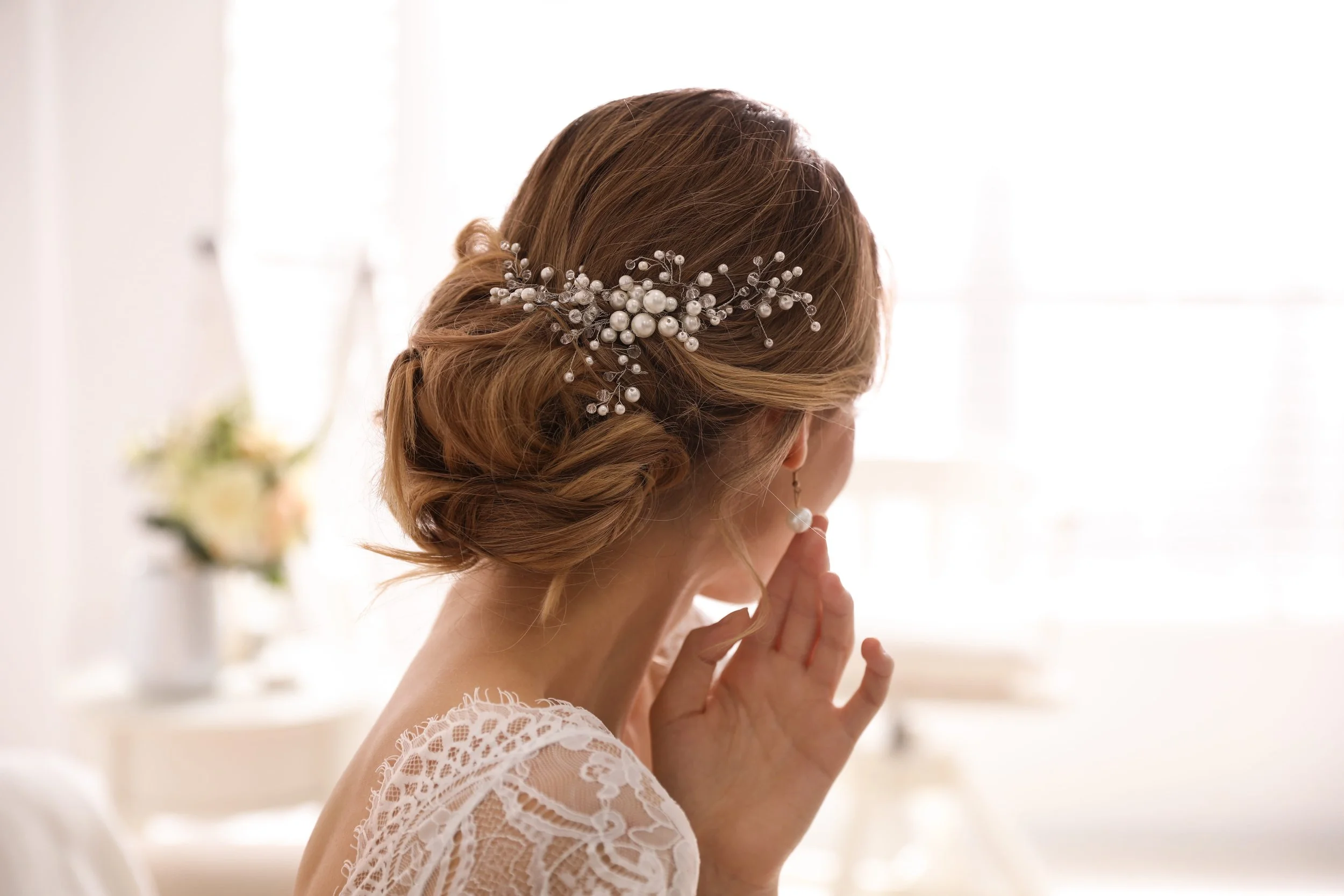A woman with styled brown hair adorned with a decorative hairpiece made of pearls and wire, wearing a white lace dress, standing inside with a bright background.